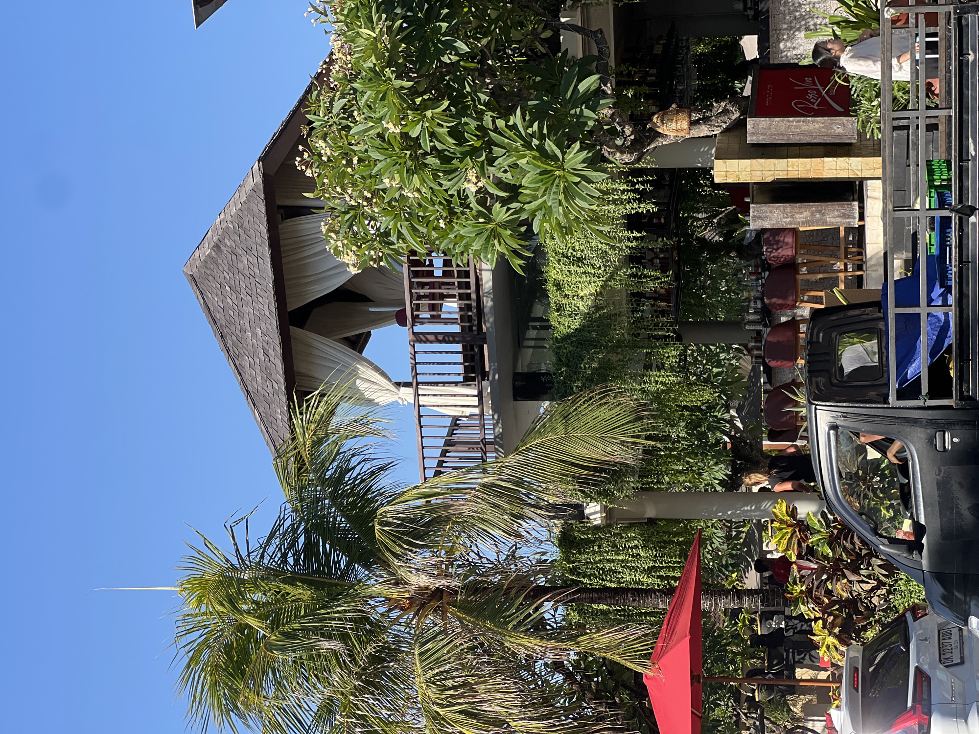 In Kuta, Indonesia, on a bright, sunny day, a multi-level building with a thatched roof structure on top is visible. Palm trees and lush green foliage surround the building. A bright red umbrella provides shade in the foreground. Several people are seen in the background, some seated at tables, others standing. A pickup truck is parked in front of the building, with a driver inside and its cargo area loaded with various items. Cars are also present on the street. The overall scene depicts a vibrant street view in a tropical location.