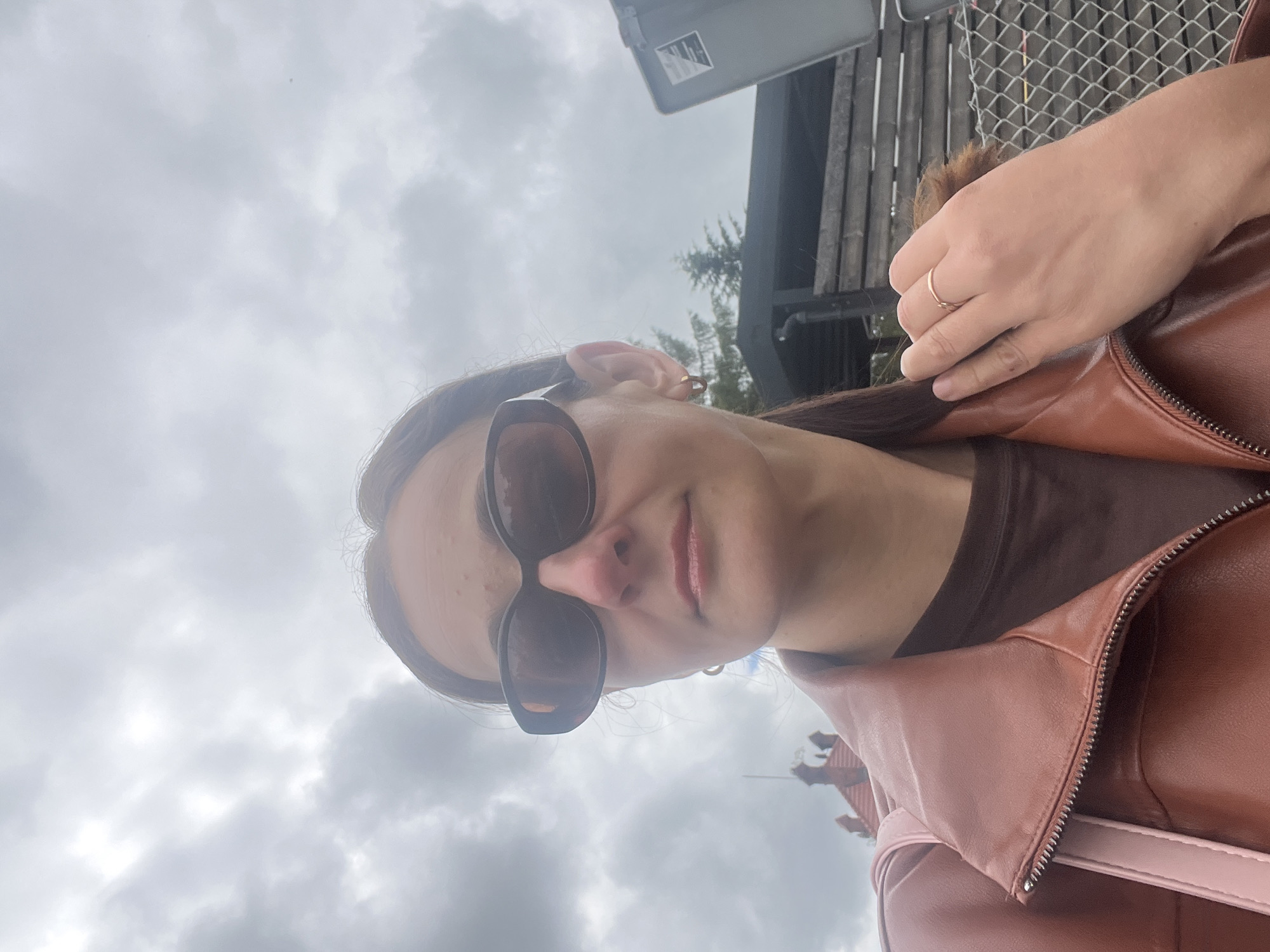 A medium shot captures a woman from the chest up, taken outdoors under a cloudy sky. She is wearing sunglasses and a brown jacket over a brown shirt. A pink bag is slung over her left shoulder. Her right hand, adorned with a ring, is resting on her shoulder. In the background, a building with a red tiled roof and a grey bin are visible. The scene is set in an urban environment, with a fence also present in the background.