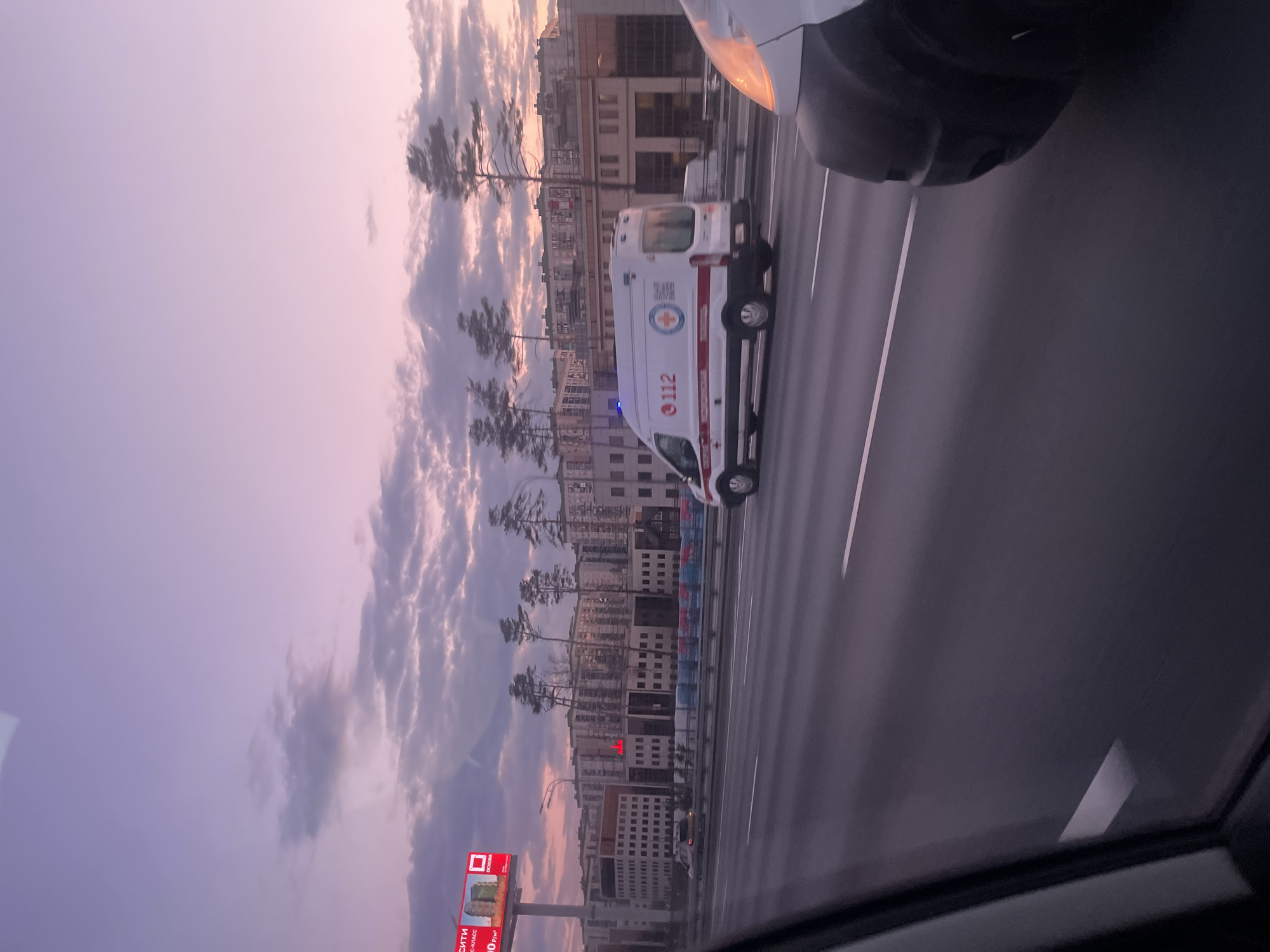 The image shows an ambulance with "112" written on it driving on a road in what appears to be Khimki, Russia, with buildings and trees in the background. The sky is cloudy with hues of pink and purple, likely during sunset or sunrise. A billboard is visible on the side of the road. The image is taken from inside a vehicle, and part of another vehicle is visible in the foreground.