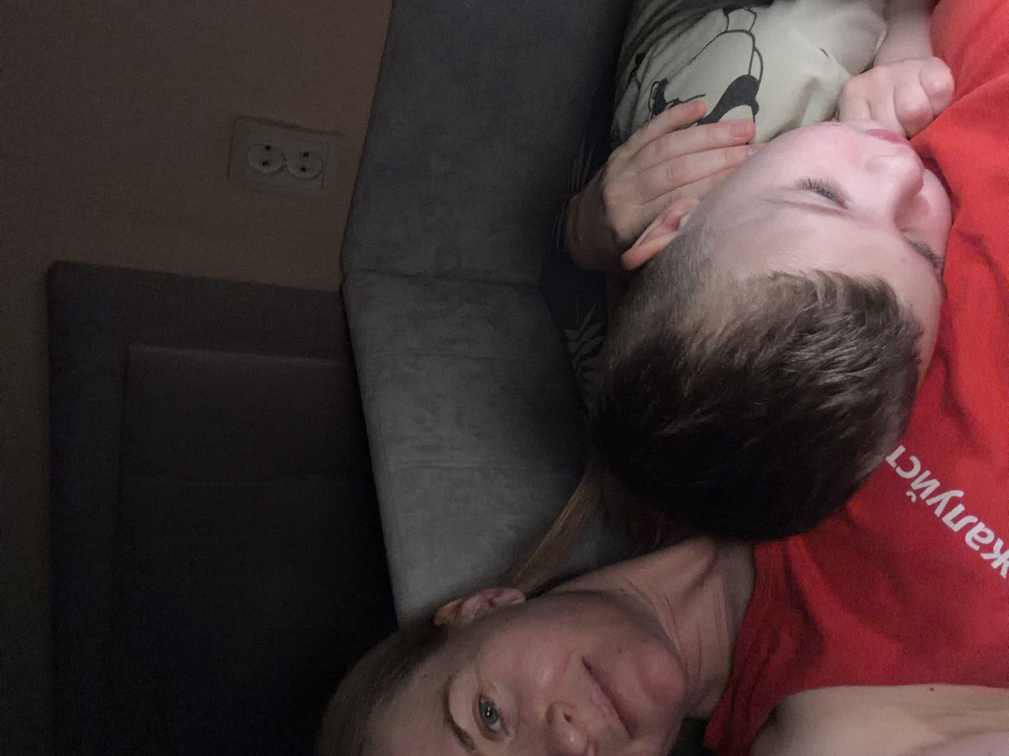 A woman and a child are lying down together in a dimly lit room. The woman is on the left, her face partially visible as she looks towards the camera with a slight smile. She is wearing a red top with white Cyrillic text that reads "Жалуйст". The child is on the right, sleeping soundly with their head resting on the woman's chest and shoulder. Their eyes are closed, and their hand is near their mouth. The background shows a dark wall, a piece of furniture, and an electrical outlet with two sockets. The scene suggests a moment of rest and connection between the woman and child. The presence of Cyrillic text on the woman's shirt, combined with the commonality of such domestic scenes, suggests this could be a typical home environment in Khabarovsk, Russia.