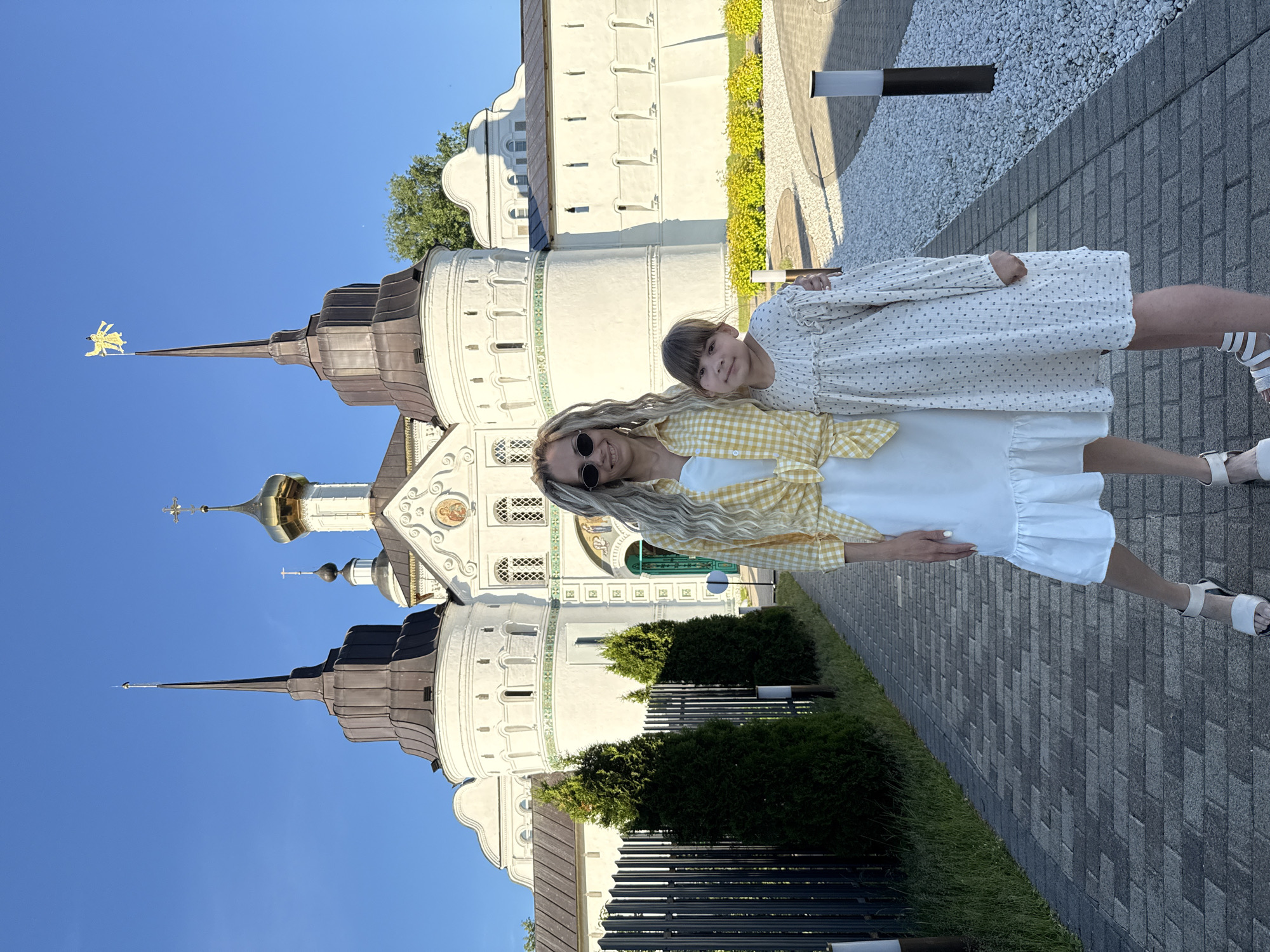An adult woman and a young girl are standing on a paved walkway, posing in front of the Vysotsky Monastery in Serpukhov, Russia, located near the city of Dumino. The woman, on the left, has long, light blonde hair styled in dreadlocks or similar braids, wears round sunglasses, a white dress with a ruffled hem, a yellow and white gingham plaid shirt tied at her waist, and white sandals. She smiles towards the viewer, with her right arm around the young girl's left shoulder. The girl, positioned to the right, has brown, shoulder-length hair with bangs and wears a long-sleeved white dress with a small dark polka dot pattern and light-colored sandals. She also looks towards the viewer with a slight smile.
The background features the distinctive white architecture of the Vysotsky Monastery, characterized by multiple towers and domes under a clear blue sky. A central building has a golden onion dome topped with a cross, flanked by two taller, conical spires with dark roofing. The spire on the right is topped with a golden angel figure. The white walls of the monastery are adorned with decorative elements, including arched windows, green accents, and a central arched entrance with a green door and what appears to be a religious fresco or mosaic above it.
The paved walkway in the foreground is made of rectangular grey pavers. To the left of the walkway, there is a dark metal fence and several neatly trimmed, conical evergreen bushes. To the right, the walkway borders an area covered in white gravel and lined with low, bright green-yellow bushes, with a dark pole-style light fixture visible. The scene is brightly lit by natural sunlight, suggesting a clear day.