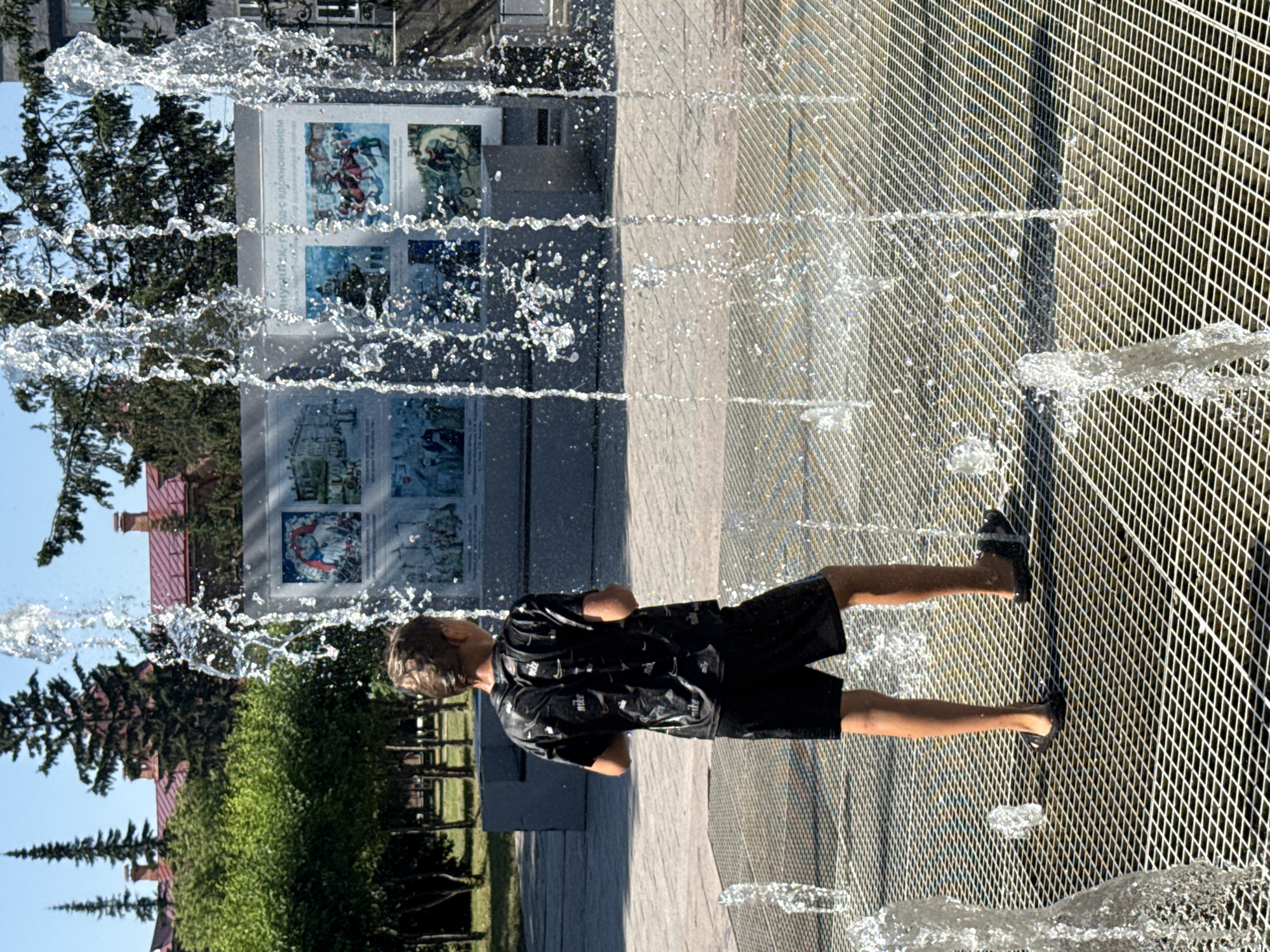 A young boy stands on a metal grate in front of a fountain in the city of Minusinsk, Russia. He is wearing a black t-shirt with a repeating Nike logo and black shorts, both wet from the water. The boy is looking towards the right of the frame, away from the camera, with his hands by his sides. Water from the fountain sprays around him, creating a dynamic and refreshing scene. Behind the boy, a large mural depicting historical scenes is visible on a wall, with trees and buildings in the background. The sun is shining, casting shadows and illuminating the water droplets. The environment appears to be a public space, likely a park or town square, given the presence of the fountain and mural.