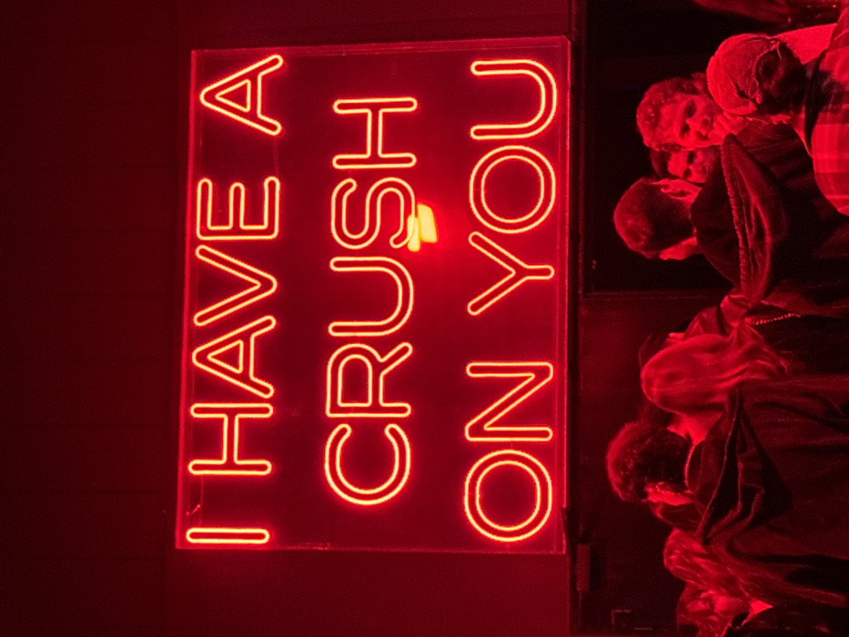 The image displays an interior scene characterized by pervasive red illumination, centered on a large, rectangular neon sign. The sign, mounted on a dark surface in the upper portion of the frame, emits bright red light, spelling out the phrase "I HAVE A CRUSH ON YOU" in capital letters. Below the sign, approximately 10-15 individuals are gathered in a dense crowd, with their heads and upper torsos visible. The strong red light casts deep shadows and defines contours. Most individuals face away from the viewer or are in profile, standing and interacting within the group. One person on the far right exhibits curly hair and a visible smile while looking left. Another individual on the immediate right wears a patterned top and a head covering. The setting is consistent with a nightlife or event venue. This specific installation and environment are located at Base Milano, Via Bergognone, 34, 20144 Milan, Italy.