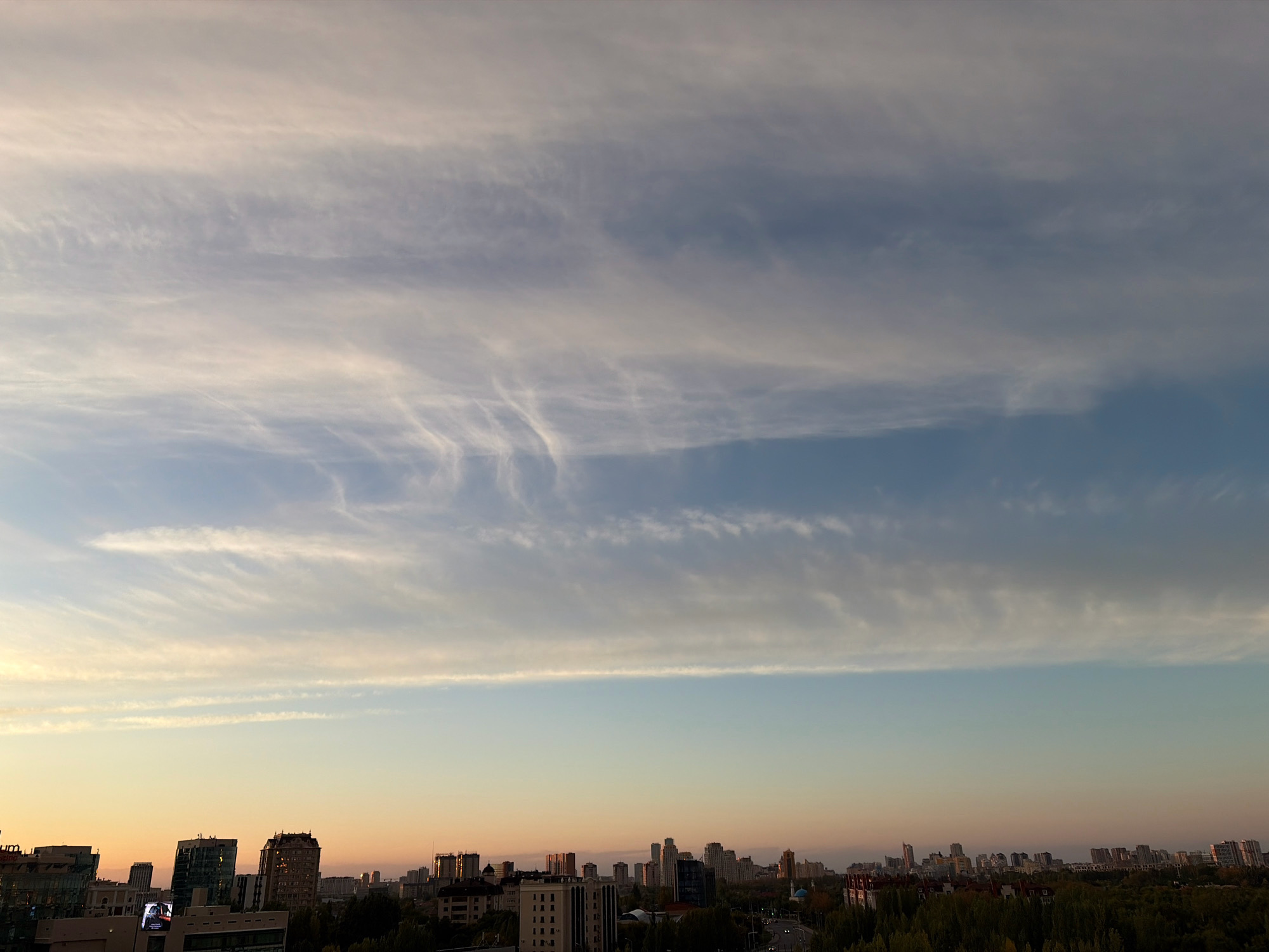 The image captures a panoramic view of the city of Astana, Kazakhstan, at what appears to be either sunrise or sunset. The sky is dominated by wispy, elongated clouds that stretch across the frame, displaying varying shades of white, gray, and light blue. Below the expansive sky, the city skyline is visible, characterized by a mix of modern high-rise buildings and lower-rise structures. The buildings are clustered together, with some taller glass-fronted towers reflecting the ambient light, and others featuring more traditional architectural details. The lower portions of the image reveal a dense canopy of green trees, suggesting a significant presence of parks or residential areas with landscaping. The overall atmosphere is serene, with the soft lighting indicating a transition period of the day. There are no discernible people or active engagements visible in the scene.