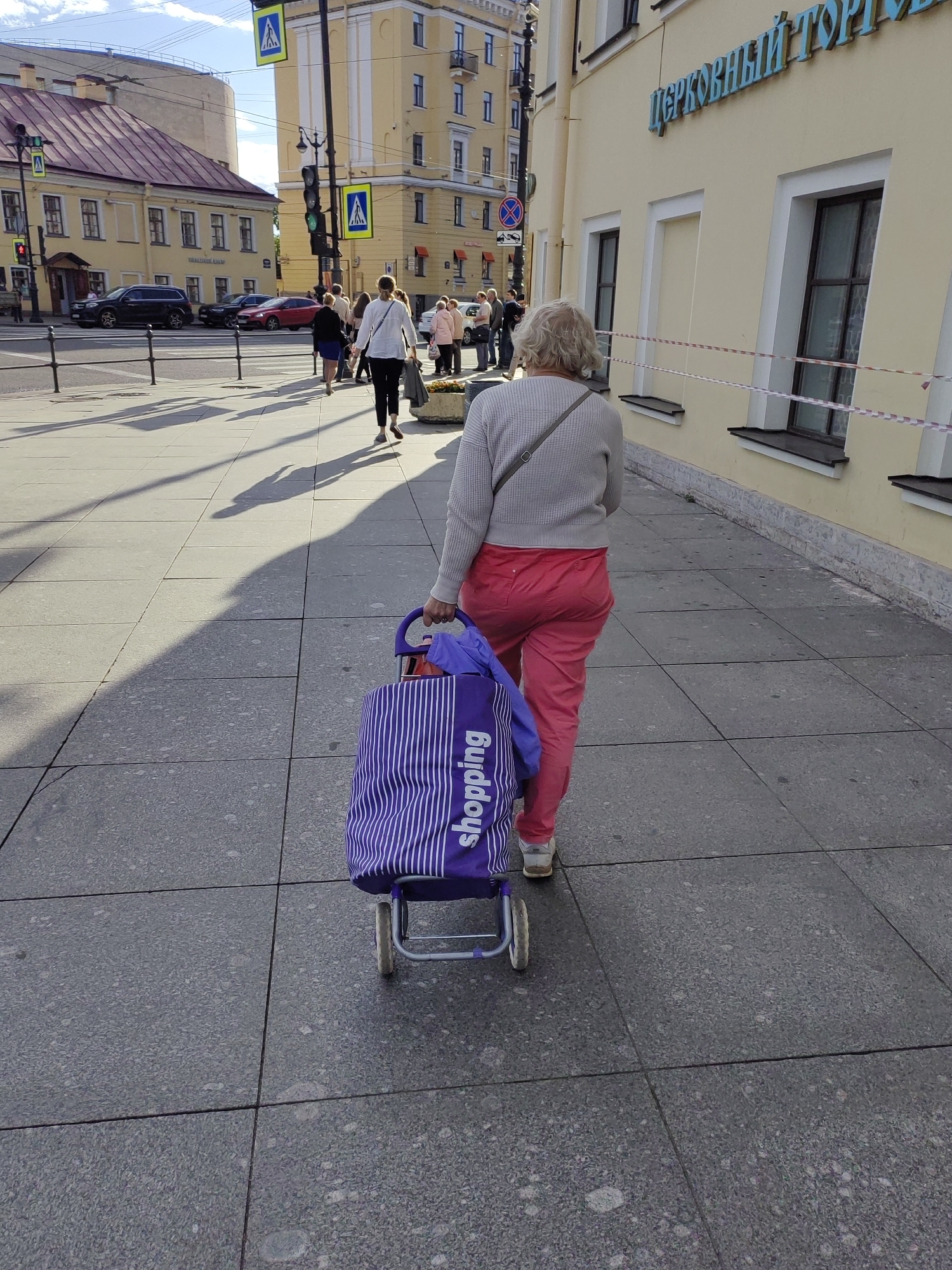 A woman walks down a city sidewalk, pulling a purple shopping cart. She is wearing red pants and a light gray sweater, with a crossbody bag slung over her shoulder. The shopping cart is striped and has the word "shopping" printed on it. The sidewalk is made of large, gray square tiles.
The background shows a street with cars and other pedestrians. On the right side of the image is a yellow building with several windows and a sign with blue Cyrillic text that reads "церковный торг" (church trade). Traffic lights are visible in the distance, and pedestrian crossing signs are posted. The sun is casting long shadows, suggesting it is either morning or late afternoon. The sky is partly cloudy. The overall scene depicts a typical urban environment on a sunny day.