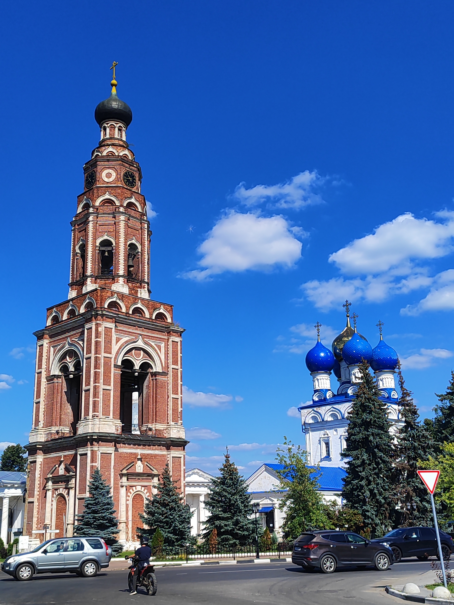 In Bronnitsy, Russia, under a bright blue sky with scattered white clouds, a detailed scene unfolds. A tall, red-brick bell tower with white detailing and arched openings stands prominently on the left. Atop the tower is a dark dome topped with a golden cross. To the right of the bell tower, a white church with blue domes and golden crosses rises against the sky. Evergreen trees are visible between the bell tower and the church. In the foreground, a road runs horizontally across the image. A silver SUV is driving on the left side of the road, and a person on a motorcycle is heading away from the viewer on the right side of the road. Another dark SUV is driving on the right side of the road. A red and white triangular road sign is visible on the right edge of the image.