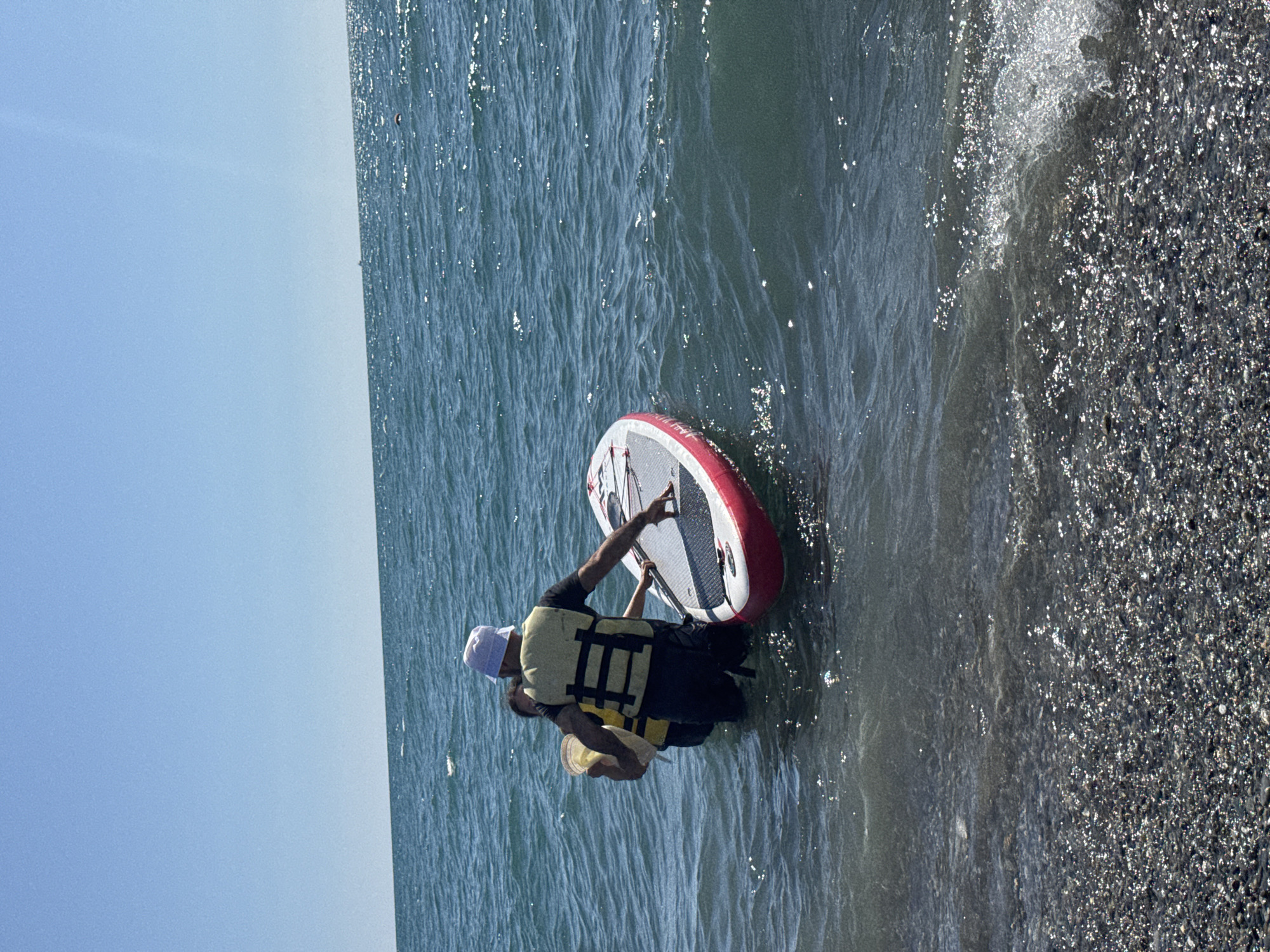 Two individuals, one wearing a white bucket hat and a yellow life vest over a dark shirt and pants, and the other person mostly obscured but also wearing a yellow life vest, are in shallow ocean water near a pebble beach. They are interacting with a stand-up paddleboard with red accents, which is partially submerged. The person in the hat appears to be steadying or adjusting the paddleboard, while the other person is positioned behind them, holding a yellow bucket. The water is a deep blue-green, with sunlight reflecting off its surface. The sky is clear and bright blue. The scene takes place on the coast of Sochi, Russia.