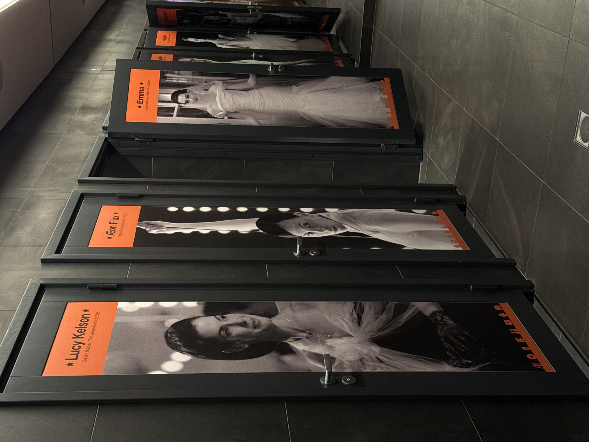 This is a low-angle, eye-level shot of a display of framed movie posters or portraits, arranged vertically in a hallway or corridor. The frames are black, and each contains a black and white image of a woman, likely an actress, accompanied by an orange label with text. The floor is tiled in a dark grey stone. The display appears to be part of an exhibition.
The visible text on the orange labels identifies the subjects and associated films:
- The top visible label reads "*Emma *" followed by "Anne Hathaway | Bride Wars | 2009". The image shows a woman in a white, flowing dress.
- The next label down reads "*Æon Flux*" followed by "Charlize Theron | Æon Flux | 2005". The image is partially obscured but shows a person's face.
- The bottom visible label reads "*Lucy Kelson *" followed by "Sandra Bullock | Two Weeks Notice | 2002". The image shows a woman's face and upper body.
The lighting is artificial, and the overall mood is somewhat somber due to the dark colors and the angle of the shot. The environment suggests an indoor public space like a cinema, gallery, or museum. The perspective and the way the frames are mounted suggest they might be doors to private viewing rooms or exhibits. The lighting creates shadows that emphasize the texture of the floor tiles.