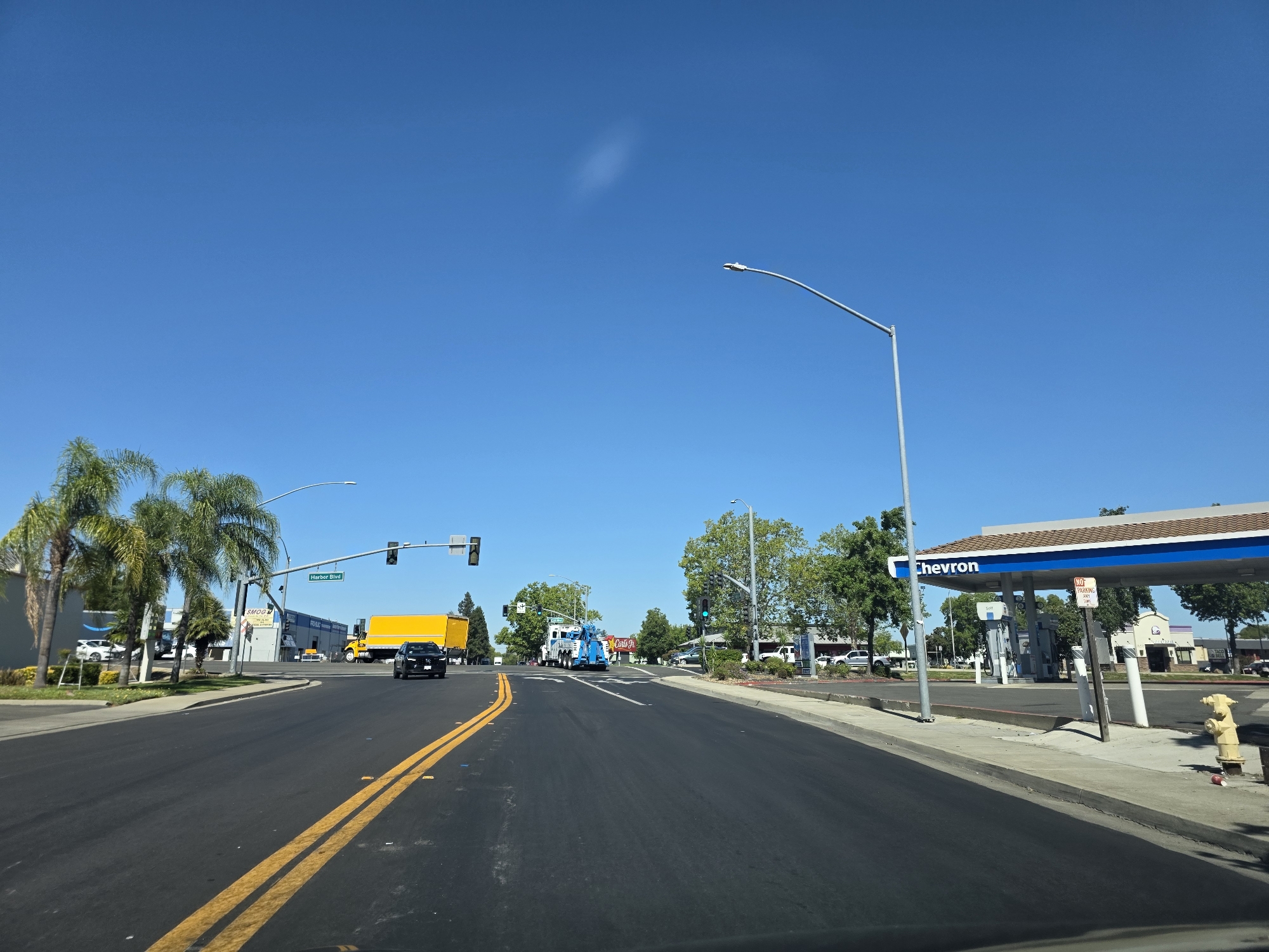 This media file captures a bright, sunny day in West Sacramento, California, likely in the late morning or early afternoon given the strong sunlight and clear blue sky. The scene is a wide, asphalt road with double yellow lines indicating no passing. On the left side of the road, palm trees line the sidewalk in front of a commercial building with an "Amazon" logo visible. Further down the road on the left, a yellow truck is stopped at an intersection, with a traffic light showing green.
The right side of the road features a Chevron gas station with its distinctive blue and white canopy. Several gas pumps are visible under the canopy, and a "NO PARKING" sign is posted. Adjacent to the gas station, other commercial buildings are visible, including one with a "Taco Bell" logo. A bright yellow fire hydrant stands on the sidewalk.
In the mid-ground, a blue tow truck is parked, and other vehicles are scattered in parking lots. The overall impression is a typical suburban commercial area on a clear, pleasant day. There is no indication of people actively engaged in any particular activity, but the presence of vehicles and businesses suggests normal daytime operations. The clear weather and abundant sunlight create a cheerful and ordinary atmosphere. Visible text includes "Chevron," "Amazon," "SMOG," "Harbor Blvd," and "Taco Bell."