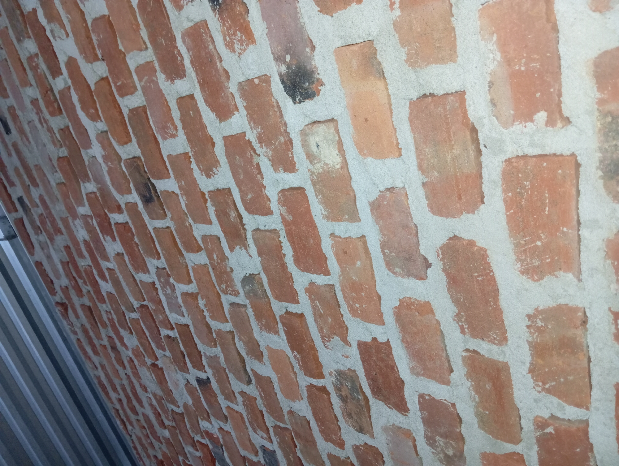 The image displays a close-up, angled view of a brick wall constructed with reddish-orange bricks and light gray mortar. The bricks are laid in a staggered pattern, and the mortar is visible between them, creating a textured surface. Some bricks show signs of wear and discoloration, with darker patches and lighter smudges. The wall appears to be an exterior or interior surface. Above the brickwork, a section of corrugated metal roofing or siding is visible, reflecting light and exhibiting a metallic sheen. The lighting suggests an indoor setting or an overcast day, with shadows cast by the bricks. The image does not contain any people or specific identifiable landmarks within Rejosari, Indonesia.