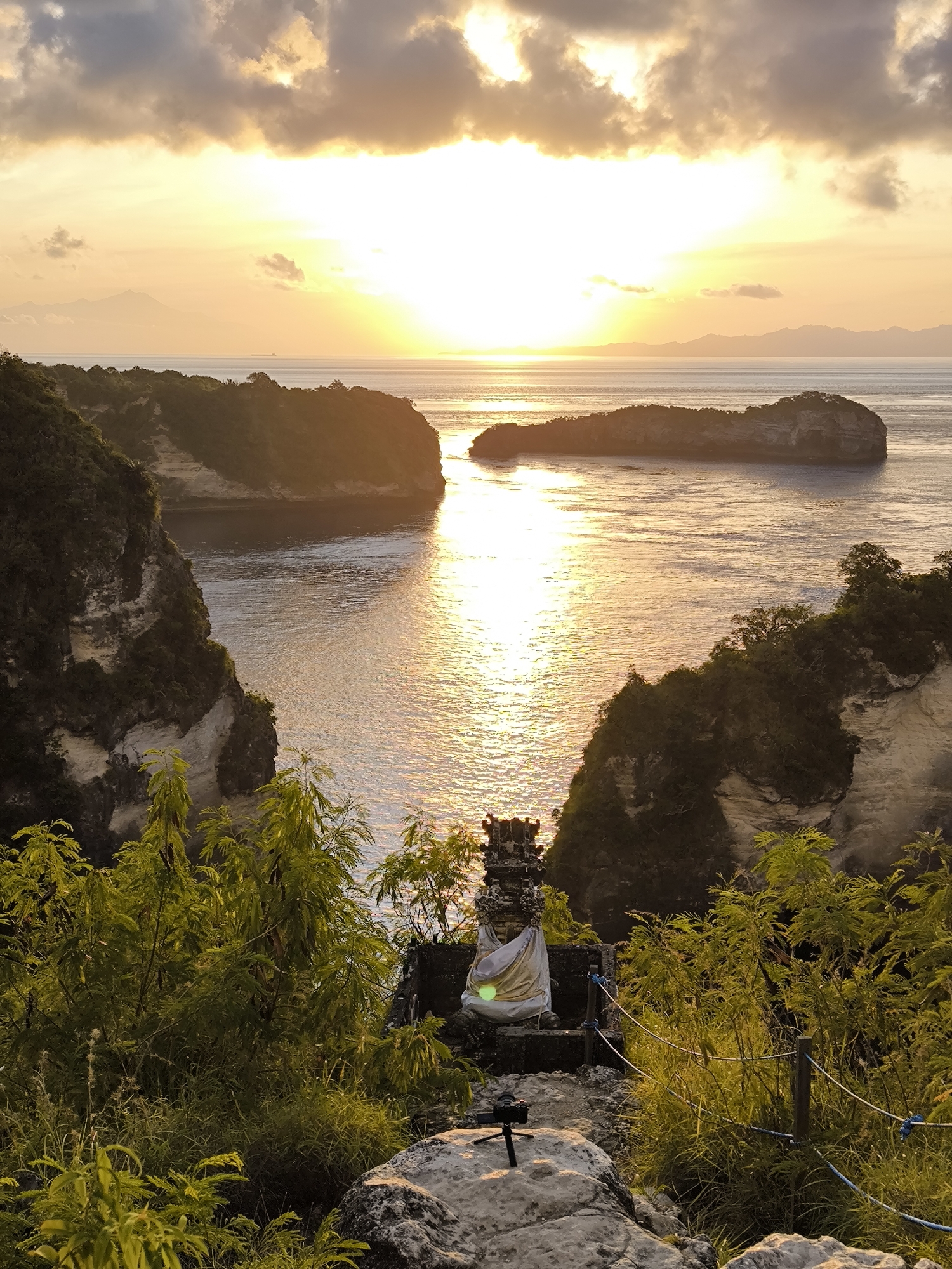A high-angle view captures a sunset scene in Nusa Penida, Indonesia. The sun's glare reflects brightly on the water, nestled between rocky cliffs and a distant island. In the foreground, vegetation frames a statue adorned with a white cloth. A small camera tripod rests on a rock beside the statue. A rope and post barrier guides a path towards the statue. Clouds fill the upper portion of the sky, tinted with the warm glow of the setting sun.