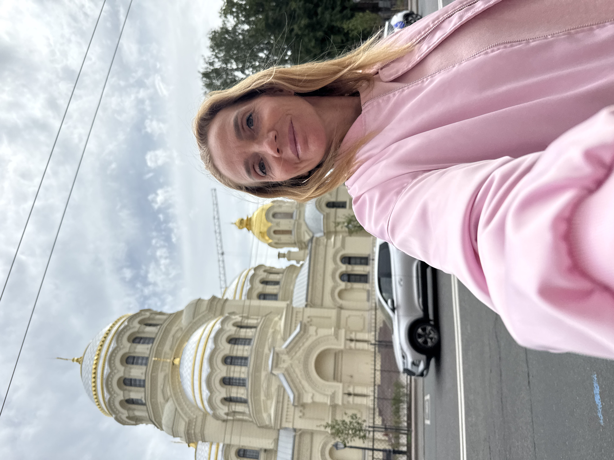 A woman with blonde hair, wearing a pink jacket and pink shirt, is taking a selfie in front of the Naval Cathedral of Saint Nicholas in Kronstadt, Saint Petersburg, Russia. The cathedral, a prominent landmark with white walls and gold domes, is visible in the background. A silver SUV is driving on the street in front of the cathedral. The sky is overcast.