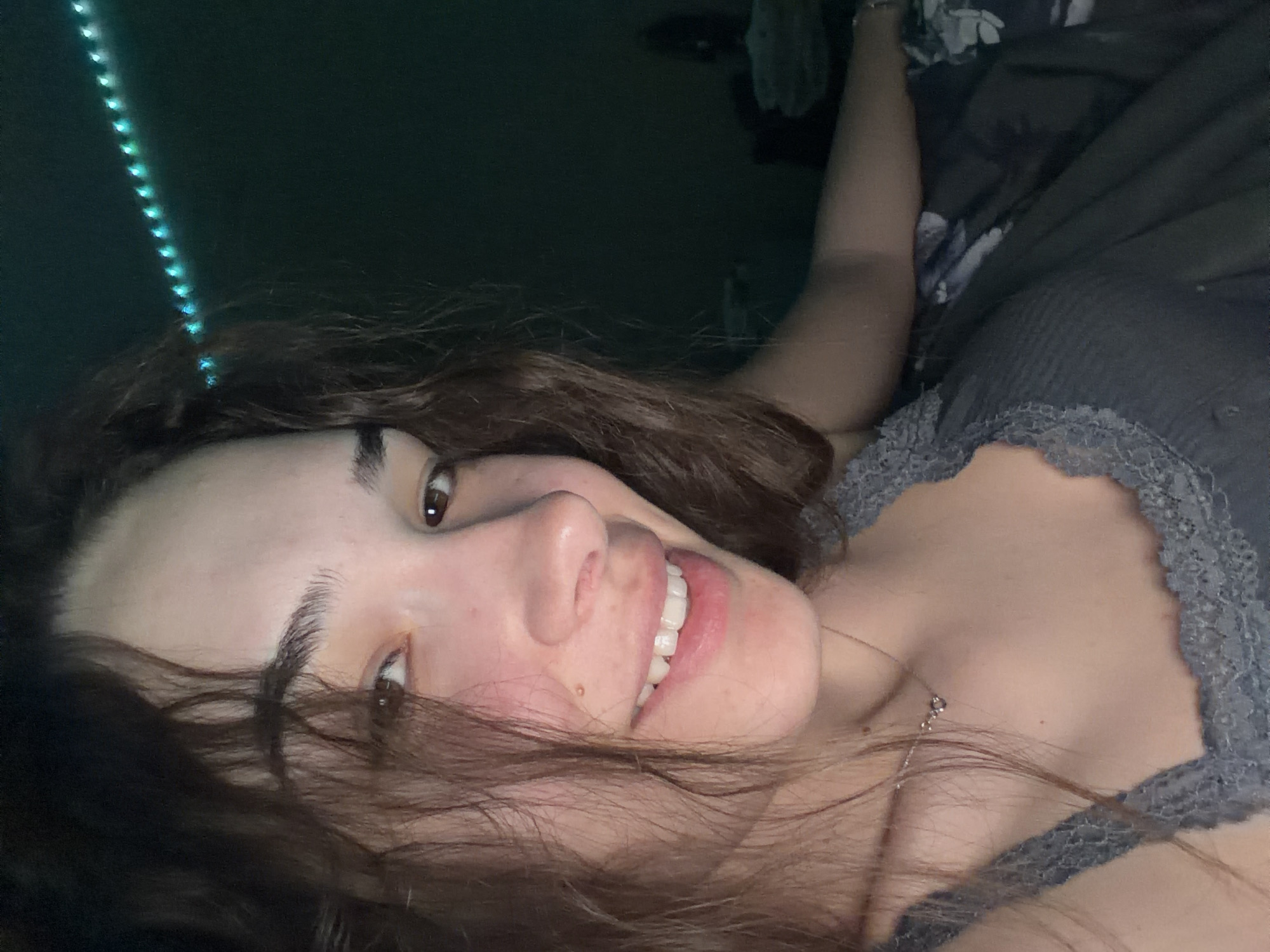 The image is a rotated, close-up portrait of a young woman, appearing to lie on her side, looking directly at the camera. She has long, wavy brown hair that frames her face, and dark eyebrows above her brown eyes. A small mole or freckle is visible on her upper lip area. She is smiling broadly, showing her white teeth, indicating a happy and relaxed mood.
She is dressed in a grey or slate blue ribbed top with a delicate lace trim along the neckline, and a thin, silver-colored necklace with a small pendant is visible around her neck.
The setting appears to be indoors, with dim lighting. In the upper left corner, a vertical string of glowing blue-green LED lights adds a decorative element. To the upper right, a dark, patterned fabric, possibly bedding or a blanket, is visible, along with a portion of her arm resting on it. The composition suggests a casual, self-taken photograph. While the context of Sofia, Bulgaria, was provided, there are no visual cues within the image to confirm this specific location. No text is visible in the image.