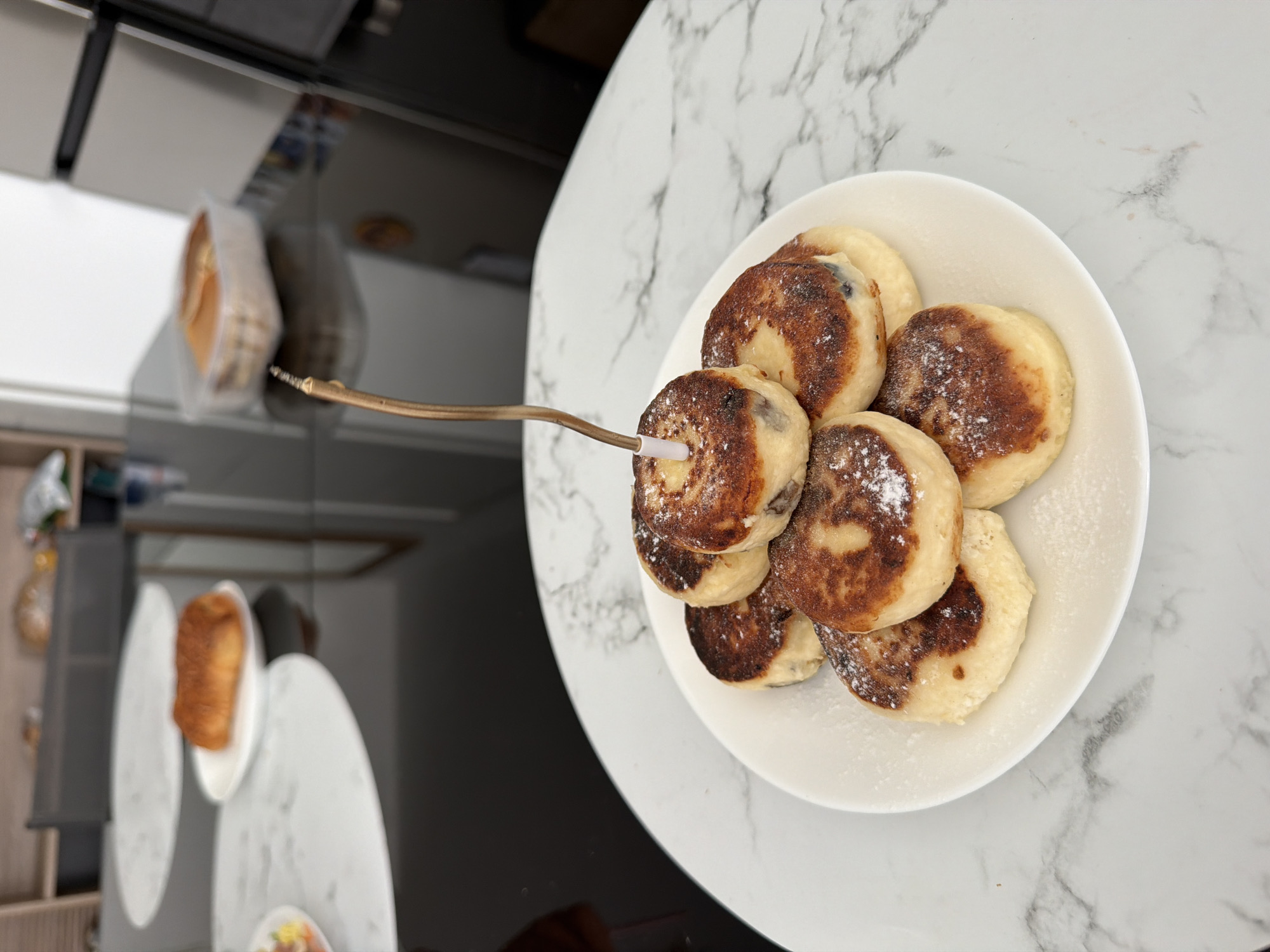 A plate of syrniki, or fried cheese pancakes, is presented on a white marble table. The syrniki are golden brown and dusted with powdered sugar. A golden candle is inserted into the top pancake, suggesting a celebration. In the background, a black reflective table is visible, holding other food items, including a croissant on a white plate and a clear plastic container with more pastries. A silver refrigerator is also in the background. The scene appears to be an indoor setting, possibly a kitchen or dining area. There are no people visible in the image. The location is not specified in the image itself, and therefore cannot be determined as Finestrat, Spain.