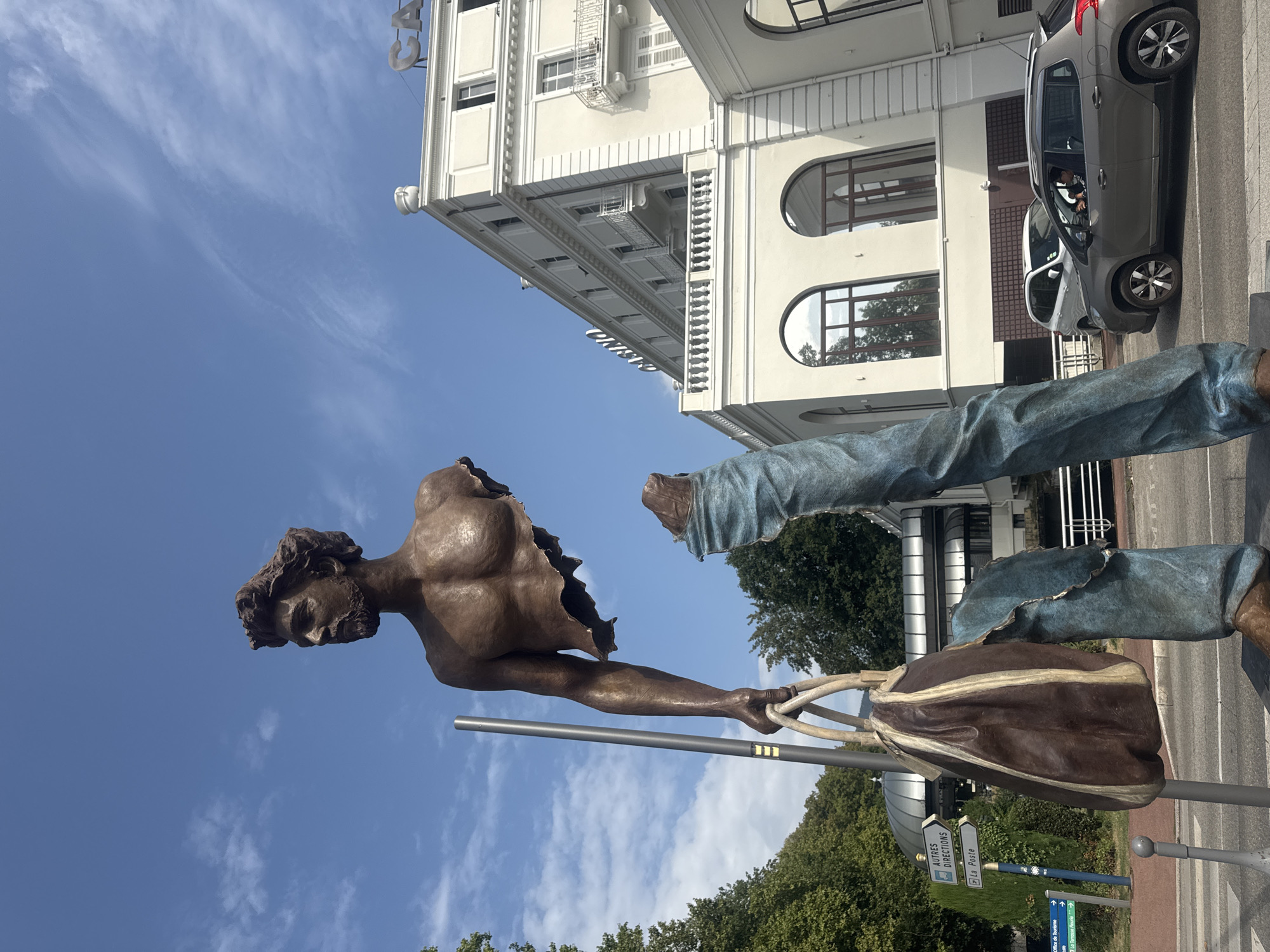 The image shows a bronze statue of a man, seemingly disembodied, with his torso and head in the upper left, and his legs in the lower right. The torso is sculpted in a realistic style, showing defined muscles and a beard. The man is holding a large, brown leather bag in his left hand. His legs are depicted in blue jeans, also in a realistic style. The statue is located outdoors in front of a white building with arched windows, which has "CASINO" written in large letters on its facade. The sky is blue with scattered clouds. There are trees in the background, and a street with cars and a traffic sign is visible in the foreground. A silver car is driving past the statue, and a white car is parked behind it. The traffic sign indicates directions to "AUTRES DIRECTIONS", "Plage", and "La Poste". The statue is situated on a street in Divonne-les-Bains, France, specifically in front of the Casino.