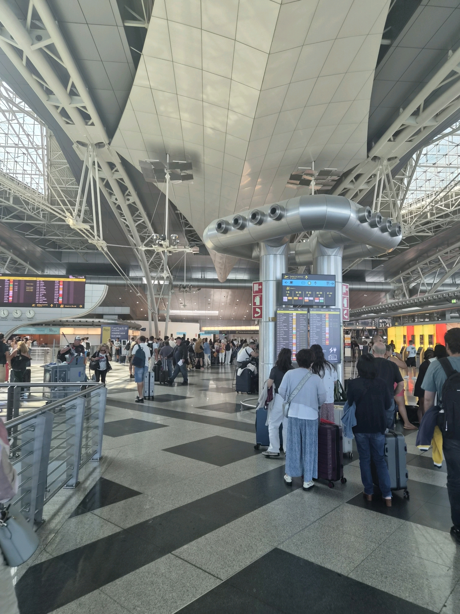 The image shows the interior of a modern airport terminal, likely located in the city of Moreira, Portugal, given the Portuguese text on the screens. The high ceiling features a complex architectural design with exposed metal beams and large, reflective panels. Natural light streams in from large windows, illuminating the spacious area.
In the foreground, several people are visible, either standing or walking with luggage. They are dressed in casual clothing, suggesting they are travelers. Many individuals are pulling wheeled suitcases, and some are standing in lines, possibly waiting for check-in or boarding.
Dominating the midground are large digital display boards showing flight information, with one screen displaying a mixture of departure times, destinations, and gate numbers in Portuguese. A prominent cylindrical structure with multiple circular openings stands near the displays, serving as a visual anchor.
The floor is made of polished granite tiles with a geometric pattern of dark and light gray squares and rectangular shapes. In the background, more travelers and check-in counters are visible, and a row of brightly colored gates or service points is present. The overall atmosphere is busy and functional, typical of an active airport environment.