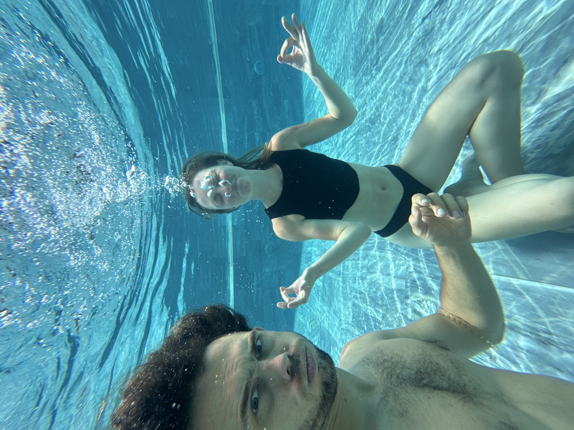 This is an underwater, wide-angle, close-up shot of two people in a swimming pool. The person on the left is a man with dark hair and a beard, looking directly at the camera with a surprised expression. He is shirtless. The person on the right is a woman with long brown hair, wearing a black bikini. She is sitting with her legs crossed, her eyes closed, and her hands in a meditative pose. Bubbles are rising around her head. The pool water is clear and blue, with sunlight filtering through from above, creating rippling patterns on the pool floor. The scene takes place in the city of Nam Hoa Lư Ward, Vietnam.