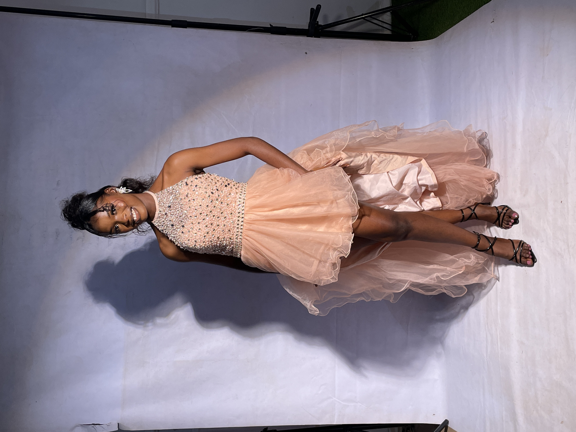 The image, taken in Barakin Akawo, Nigeria, features a young Black woman posing for a photograph in what appears to be an indoor studio setting. She is smiling broadly and looking directly at the camera, conveying a sense of joy and confidence.
The woman is adorned in an elaborate, light peach or pink formal dress. The bodice is intricately embellished with shimmering sequins or beads, featuring a high-neck design. The skirt is short and voluminous in the front, made of sheer, ruffled fabric, revealing her legs. It transitions into a longer, flowing train or overlay of similar material at the back. She wears black strappy high-heeled sandals with visible pink nail polish and distinctive earrings, possibly shaped like butterflies or flowers. Her dark hair is styled back.
The background consists of a seamless, light gray or white paper backdrop that curves from the wall to the floor. Artificial lighting is evident, casting a prominent shadow of the woman on the backdrop to her left. Visible studio equipment includes a black tripod stand and a horizontal support bar, indicating a professional photoshoot environment. A small section of green, textured material, possibly artificial grass, is visible in the top right corner. The woman's formal attire and pose suggest she is participating in a photoshoot for a special occasion or event. No text is visible in the image.