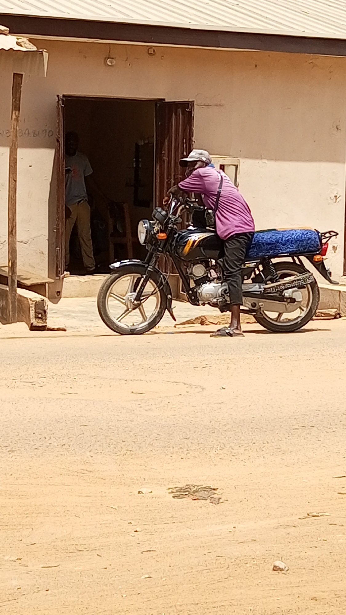 In Sabon Tasha, Nigeria, a man in a purple shirt, black pants, and sandals is leaning on a black motorcycle parked on a dirt road in front of a building. The motorcycle has a blue patterned cover on the back seat. Inside the building, a man wearing a grey shirt and light brown pants is visible.