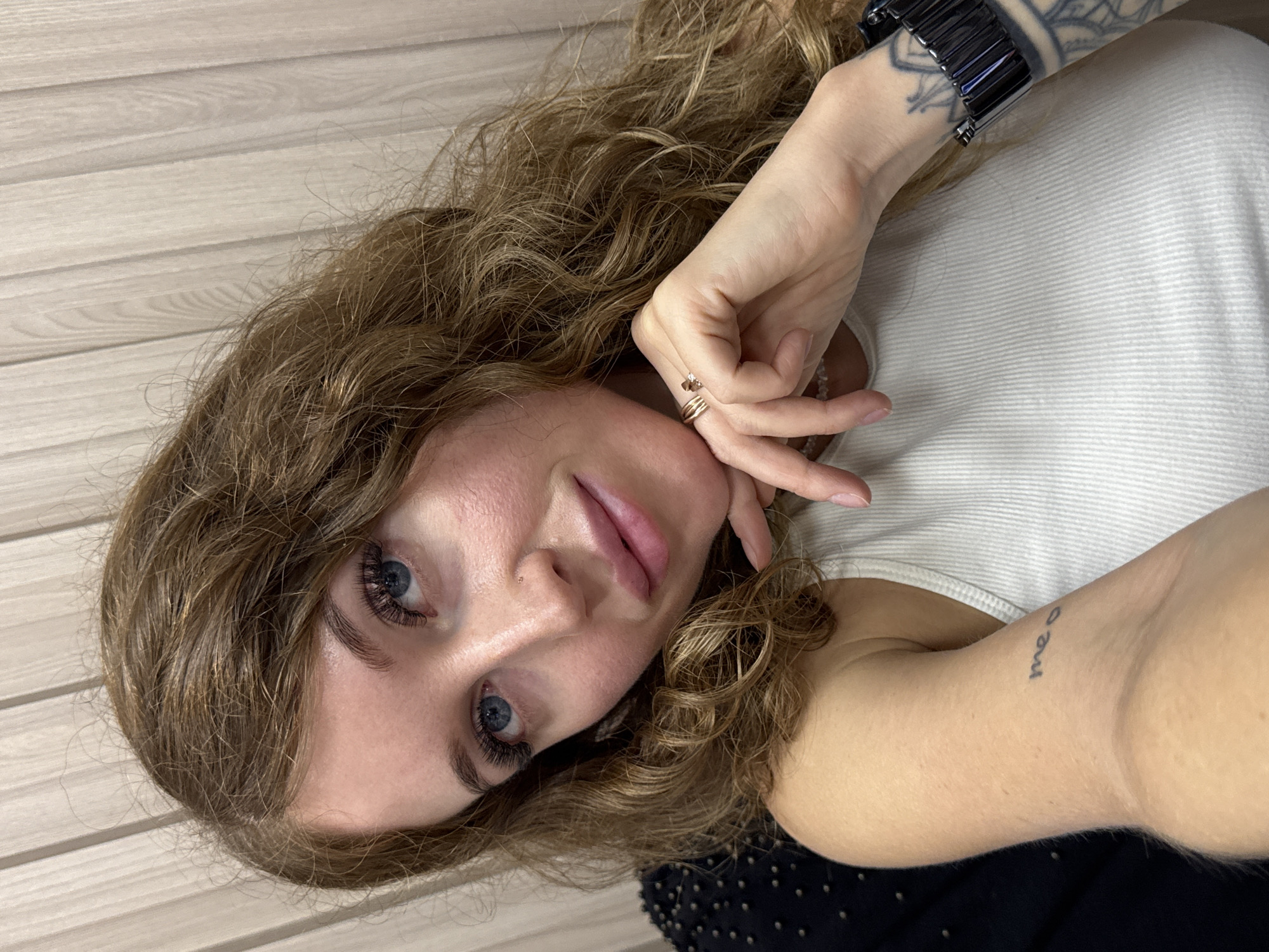 This is a close-up, selfie-style portrait of a young Caucasian woman with long, wavy brown hair and blue eyes. She is wearing a white ribbed tank top. She has on false eyelashes, rings on her left hand, and a bracelet on her right wrist. There is a small tattoo on her left forearm that appears to read "me o". Her right hand is resting on her chin. The background appears to be a wall with vertical blinds, suggesting an indoor setting. There is no visual information in the image to indicate the location is the city of Chaikovsky, Russia.
