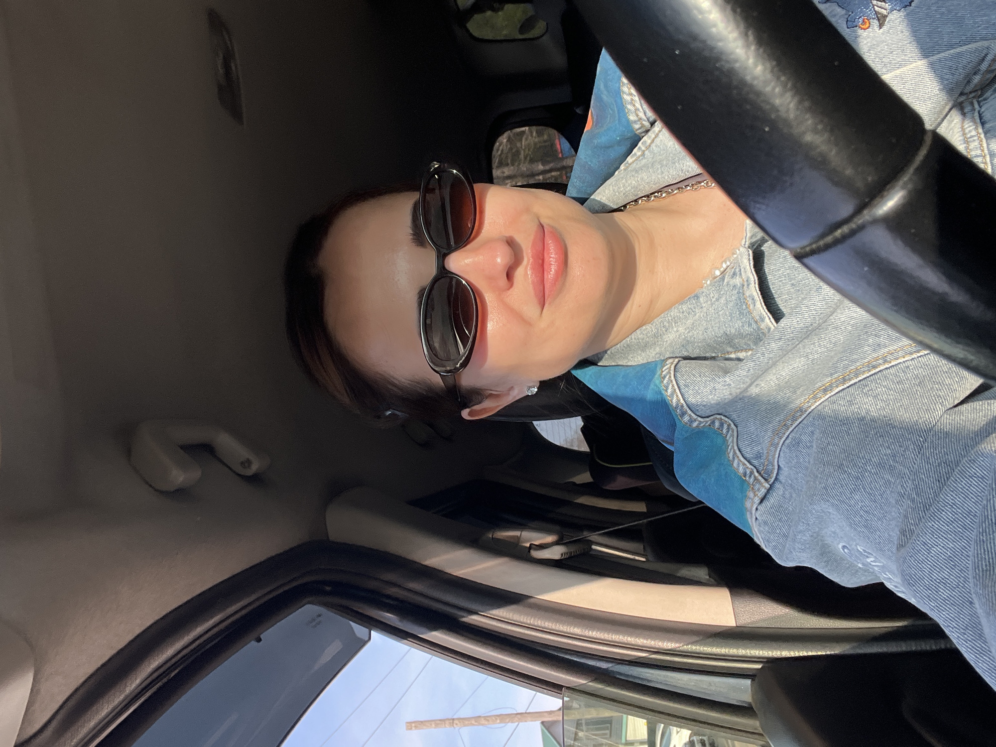 A woman wearing sunglasses and a denim jacket is seated in a car. The steering wheel is visible in the foreground. Outside the car window, there are glimpses of buildings and greenery. The location is likely in an urban area, but the specific city is not identifiable.