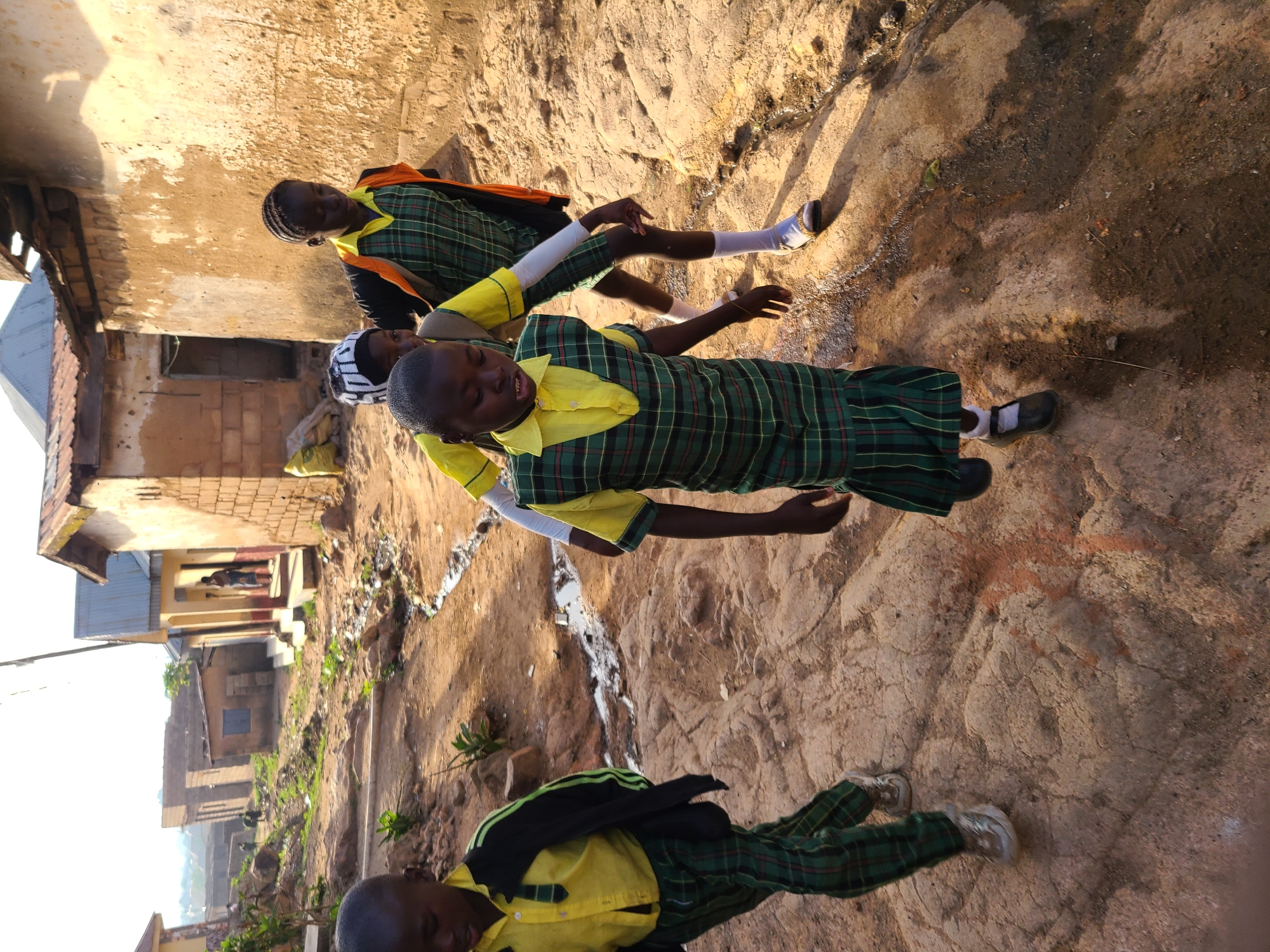 Three children are standing on a dirt path in the city of Jos, Nigeria. They are wearing green and yellow school uniforms with white socks and black shoes. One child has an orange jacket and another has a black jacket. The dirt path is cracked and uneven. Buildings are visible in the background.