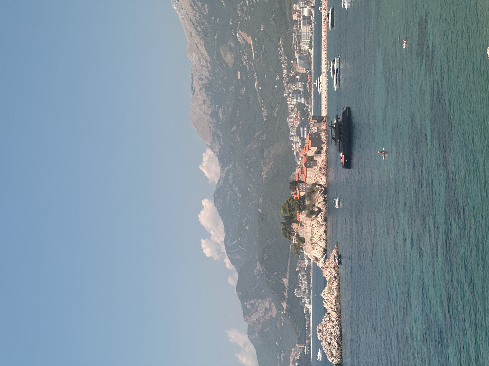 A wide shot captures the coastal town of Sveti Stefan, Montenegro, under a clear blue sky with sparse clouds. The foreground is dominated by the tranquil, turquoise waters of the Adriatic Sea. Several boats of varying sizes are scattered across the water, including a sleek, black yacht, a white motor yacht, and smaller vessels. Two individuals are visible on the water: one standing on a paddleboard, and another sitting on an inflatable float.
In the midground, the iconic Sveti Stefan island is a prominent feature, connected to the mainland by a narrow causeway. The island is adorned with red-tiled roofs of buildings nestled amongst greenery, perched atop rocky cliffs. A shoreline with buildings and a beach stretches across the background, leading to a backdrop of lush, green mountains that rise majestically into the sky. The water near the island appears clearer and shallower, revealing the seabed. The overall scene depicts a serene coastal landscape with leisure activities taking place on the water.