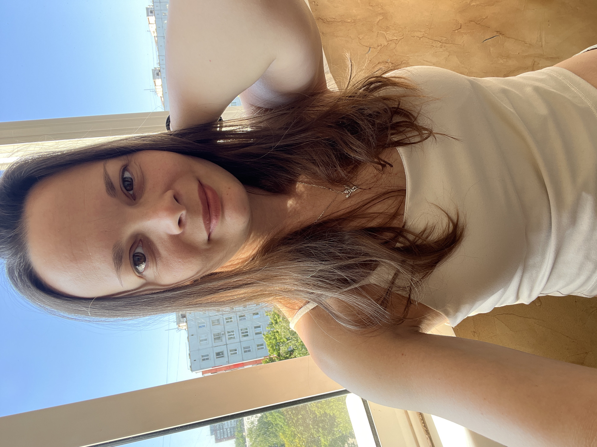 A medium-shot, selfie-style photo shows a woman in her late 20s or early 30s posing indoors in front of a window. The woman has medium-length, brown hair and fair skin. She is wearing a white tank top and a silver necklace with a small pendant. Her left arm is raised behind her head, and her right hand is partially visible in the frame.
The background is dominated by a bright blue sky and a cityscape, suggesting an urban setting. Visible buildings include a light blue apartment building with multiple windows and a red-topped building. Lush green trees are also present in the mid-ground. The interior wall to the right of the woman has a textured, orange-brown finish. The overall lighting suggests it is daytime.