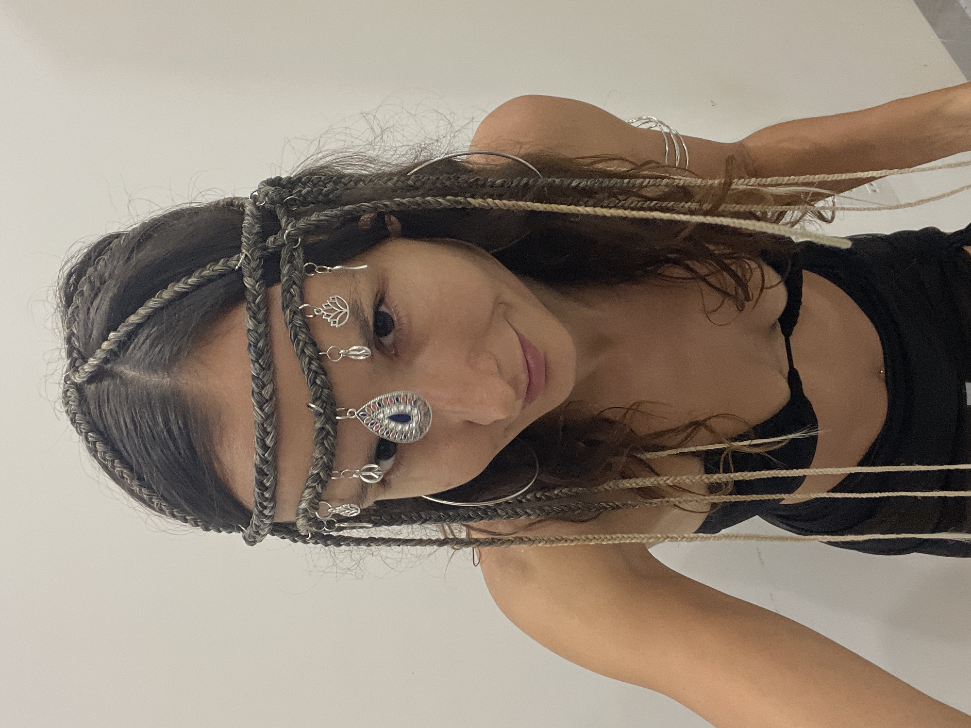 This media file shows a close-up portrait of a woman wearing elaborate braided hair adorned with silver jewelry. The jewelry includes small charms resembling lotus flowers and peace signs, as well as a larger teardrop-shaped pendant with a blue gem on her forehead. She is also wearing several thin silver bangles on her arm and a delicate silver headband. Her hair is styled in intricate braids that frame her face, with some lighter-colored strands woven throughout. She is looking towards the camera with a slight smile, her eyes visible and focused. She is wearing a black top. The background is a plain white wall, suggesting an indoor setting, possibly a studio or a room. The lighting is soft and even, indicating it's likely daytime or well-lit indoors. No text is visible in the image. The overall mood conveyed is one of adornment and perhaps a sense of artistic expression or cultural styling. The provided location context of Mũi Né, Vietnam, does not have direct visual cues within the image itself, but implies a potential cultural background or inspiration for the styling.