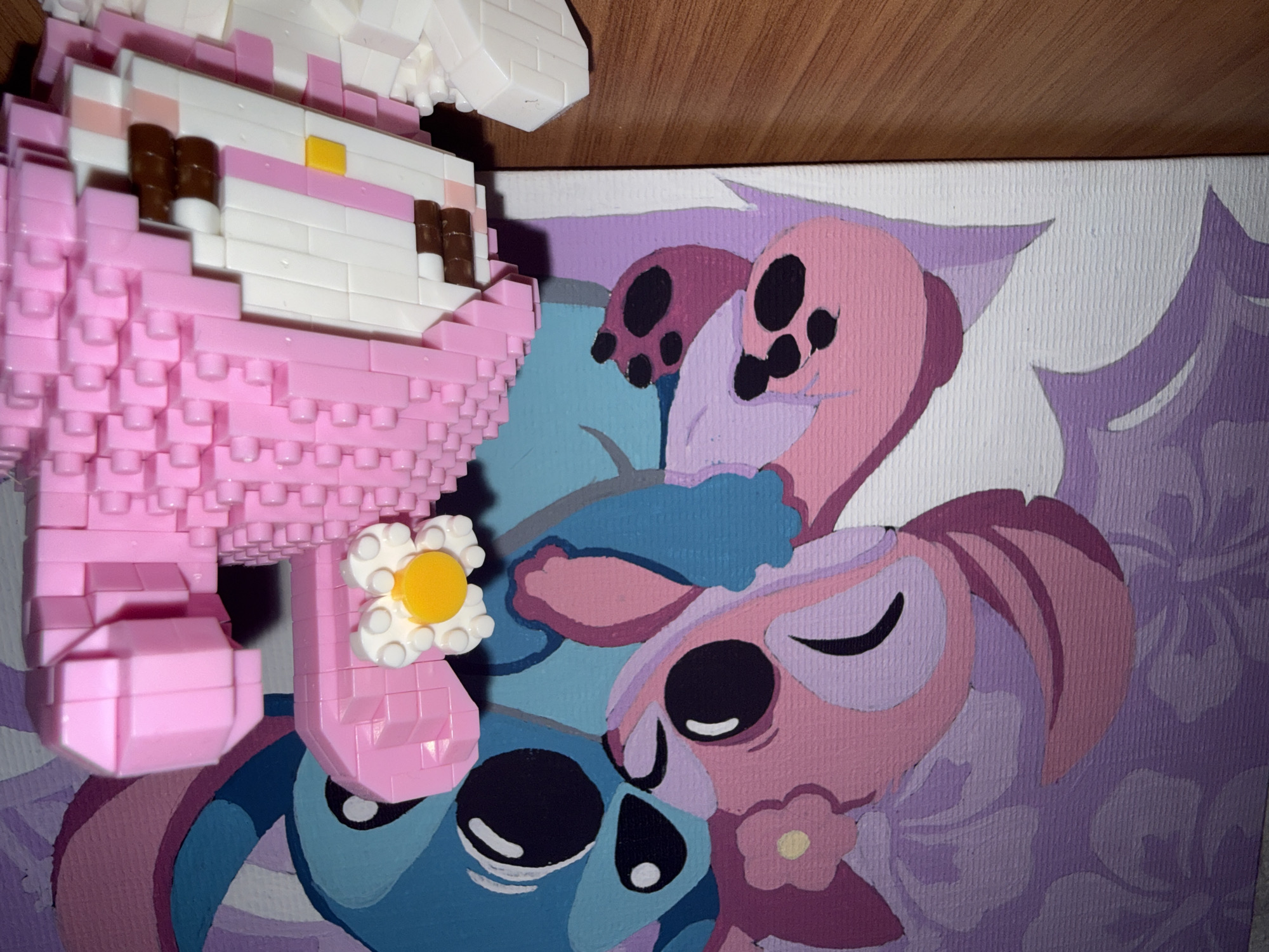 The image depicts a close-up, eye-level shot of a wooden surface with a canvas painting and a pink construction toy in the foreground. The canvas painting, positioned on the left, features cartoon characters, seemingly Stitch and Angel from Disney's Lilo & Stitch, rendered in a pastel color palette with purple floral patterns in the background. On the right, a pink construction toy, resembling a character with large pink hair and a yellow nose, is the primary focus. The toy is assembled from numerous small, interlocking pink and white plastic bricks, with brown and white details forming the eyes and mouth. A white and yellow flower element is attached to the left side of the toy. The wooden surface appears to be a table or shelf, exhibiting a visible grain pattern. There are no discernible elements in the image that can be used to determine its location within the city of Rayevskiy, Russia.