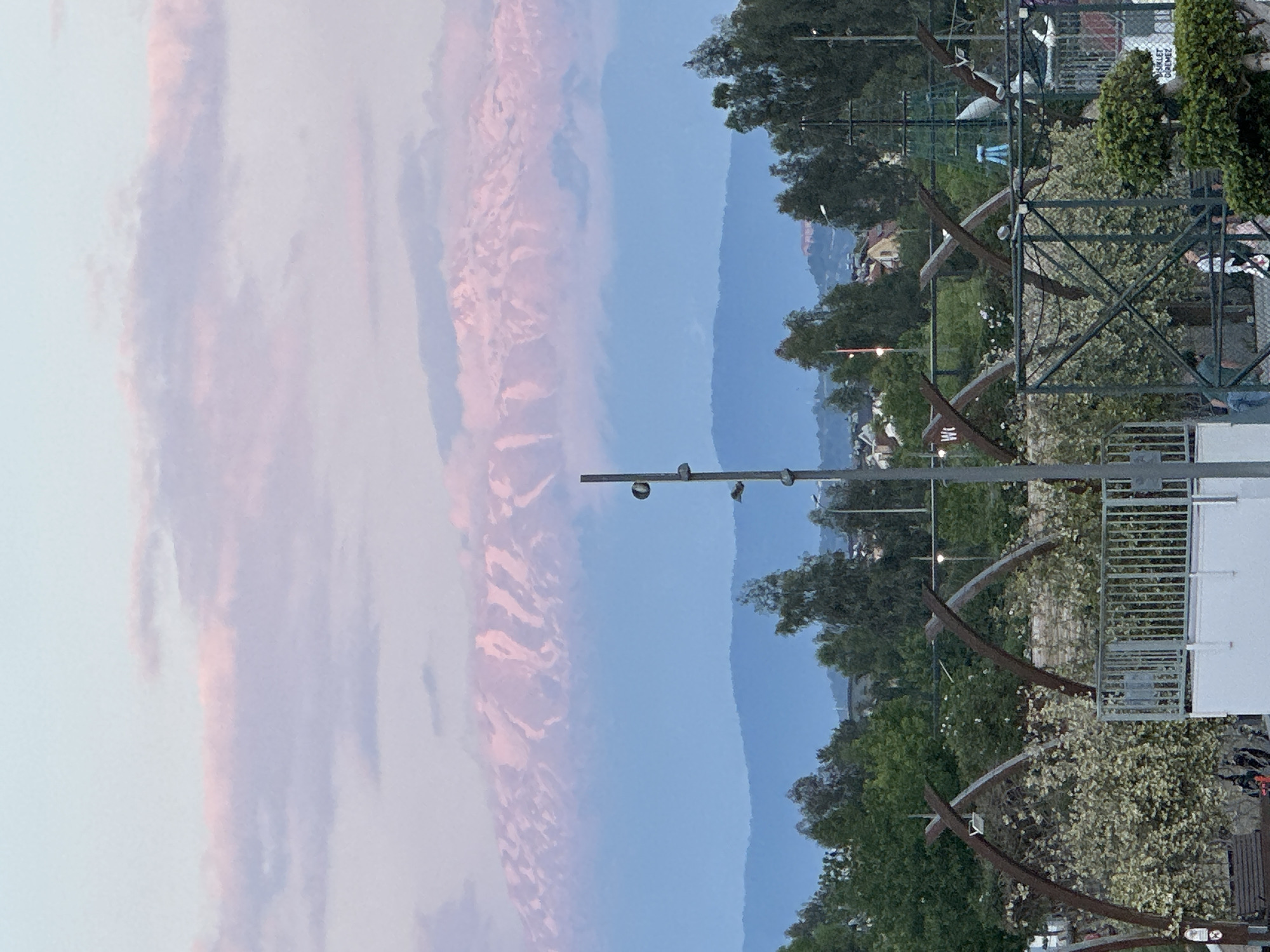 The image depicts a dusk scene from Fethiye, Turkey. The left of the image shows pink clouds against a blue sky. Moving right, a mountain range is visible. Further right is an urban landscape of buildings and trees. Streetlights and a metal sculpture or structure are in the foreground. The lighting indicates a low-light environment, likely sunset or twilight.