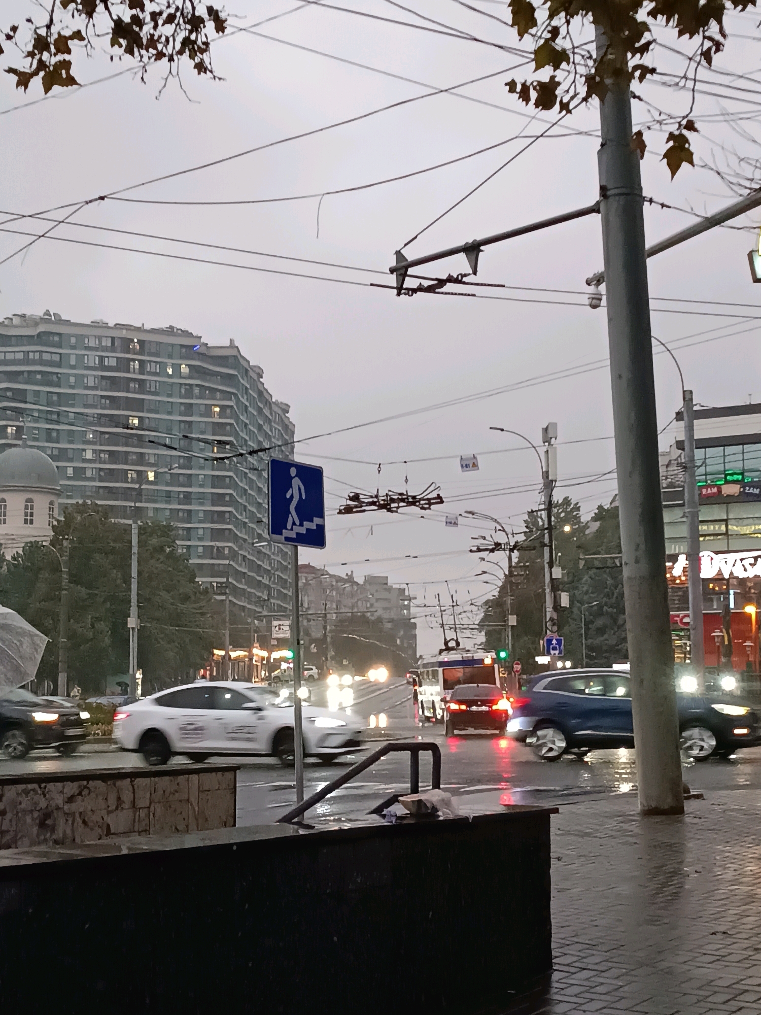 The image captures a street scene in Chișinău, Moldova, likely during overcast weather or at dusk, given the ambient light and the reflections on the wet pavement. In the foreground, a dark, rectangular structure, possibly a planter or a piece of street furniture, is visible. Beyond this, the wet asphalt street is filled with motion blur from passing vehicles. A white sedan, possibly a Tesla, is in motion, as is a dark colored SUV. Further down the street, a trolleybus and another car are also visible, their headlights and taillights creating streaks of light.
Dominating the background is a large, multi-story building with a distinctive curved facade and many windows, some of which are illuminated. To the left of this building, a smaller, older structure with a domed roof is partially visible. Overhead, a complex network of wires, characteristic of a trolleybus system, crisscrosses the sky. A traffic sign indicating a pedestrian crossing with stairs is mounted on a pole in the middle ground. The overall atmosphere is one of urban movement under diffused light, with the wet conditions suggesting recent or ongoing precipitation.