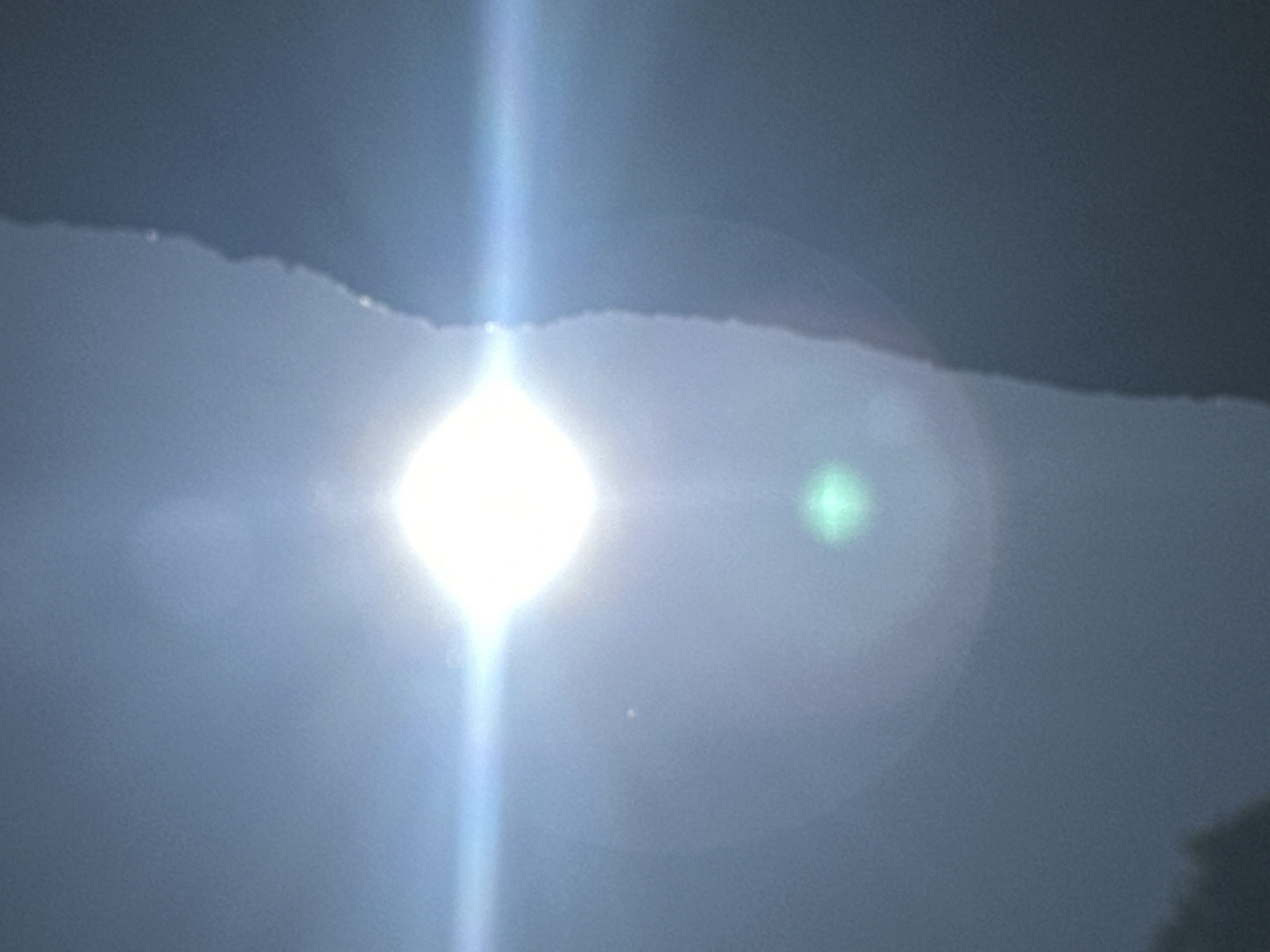 The image depicts a bright sun with lens flare, partially obscured by a dark, irregular object that appears to be a tree or a building edge. The background is a hazy blue sky. The sun's rays create a prominent horizontal light streak across the image. There is a faint green circular lens flare present below the sun. The bottom left of the image shows a dark silhouette of foliage. The overall scene is dominated by the bright light of the sun and the contrast with the dark foreground element. There is no identifiable location in Moscow, Russia, visible in this image.