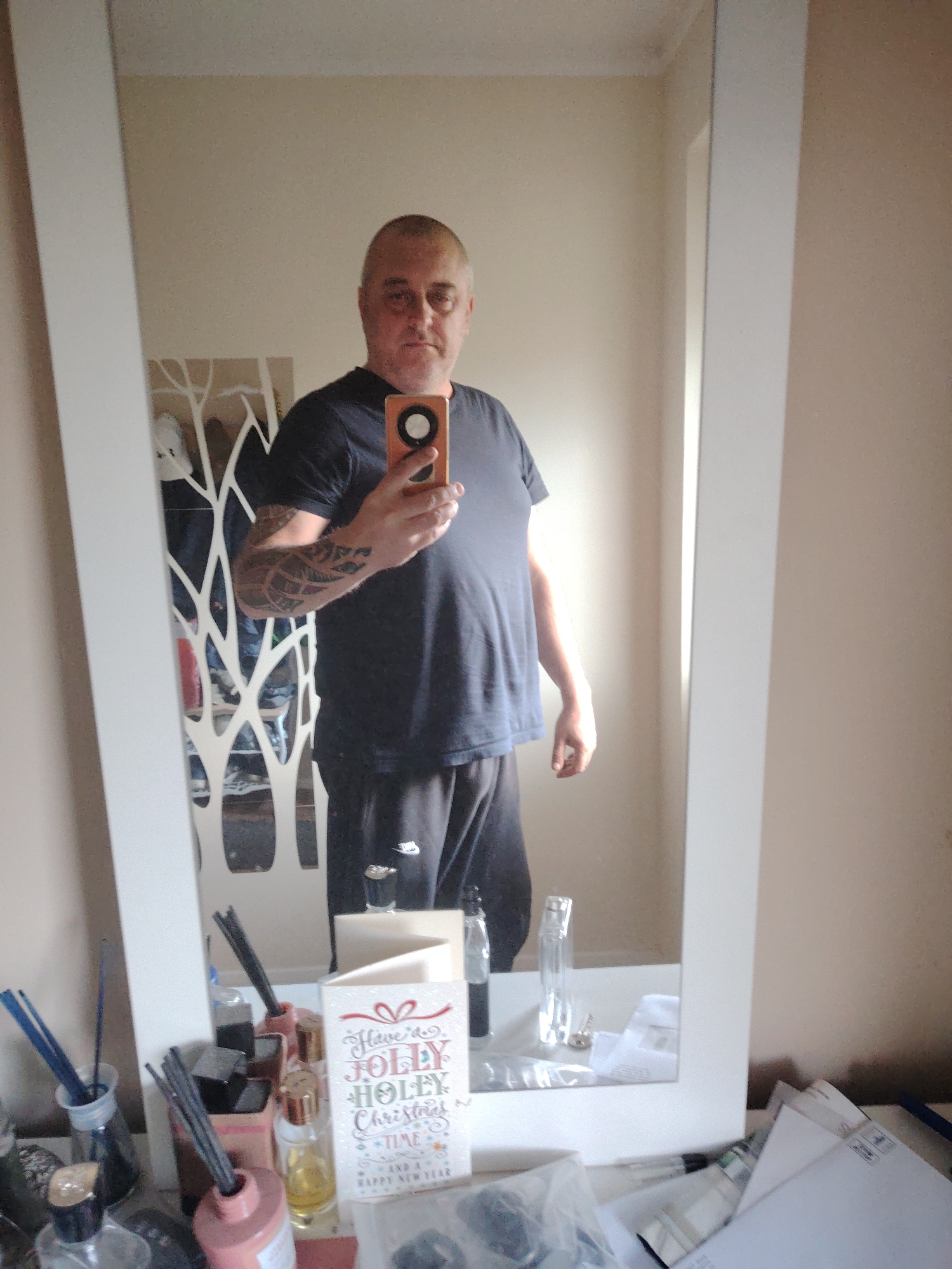A man is taking a mirror selfie indoors. He is captured from approximately the waist up, standing before a full-length mirror with a white frame. The man appears to be middle-aged, with a shaved head and a neutral facial expression. He is dressed in a dark, short-sleeved t-shirt and dark trousers. Distinct tattoos are visible on his left arm (appearing on the right in the reflection). He holds a smartphone with an orange case in his right hand (appearing on the left in the mirror) to take the photograph.
The setting is an interior room, likely a bedroom or dressing area within a home, indicated by the personal items and the full-length mirror. The walls are a plain, light beige or cream color. Reflected in the mirror behind the man is a decorative white panel featuring an abstract, branching design. Further back, partially obscured, are shelves or furniture with various items, including what appears to be footwear.
The base of the mirror rests on a surface, possibly a dresser or shelf, which is occupied by numerous objects. On the left side, several diffusers with dark reeds are placed in bottles of varying designs, some clear, some tinted pink or reddish. Smaller bottles, possibly containing perfume or essential oils, are also present. A white greeting card stands prominently in the center foreground, displaying the text: "Have a JOLLY HOLLY Christmas TIME AND A HAPPY NEW YEAR". To the right of the card, additional clear bottles or vases and a stack of white papers or envelopes are visible. The lighting is soft and ambient, suggesting an indoor setting in Huntingdonshire, United Kingdom.