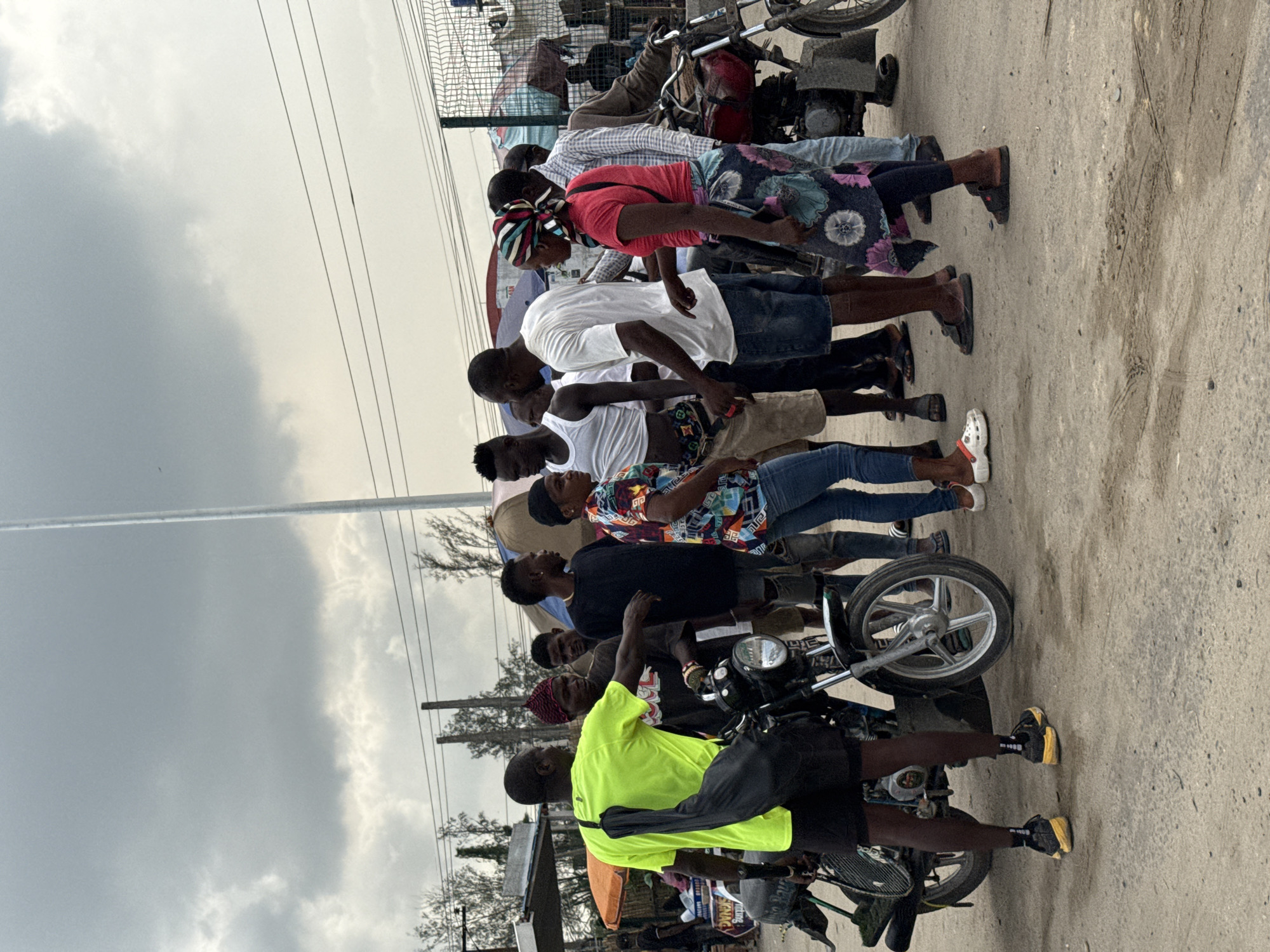 This image depicts a daytime street scene in what appears to be an urban or semi-urban environment, identified as Lekki, Nigeria. The sky is overcast, suggesting cloudy weather conditions.
A group of about a dozen people, predominantly men, are gathered on the side of a road that features both dirt and paved surfaces. They are standing or possibly waiting, with several motorcycles positioned amongst them and on the road. The presence of multiple motorcycles suggests the setting may be a common pickup point for motorcycle taxis (okadas) or a gathering spot for riders.
The individuals are dressed casually in everyday wear, including t-shirts, button-down shirts, jeans, and shorts. One man in the foreground stands out in a bright, neon yellow-green long-sleeved shirt, black shorts, and black sneakers with yellow soles, with a black bag slung over his shoulder. Other visible footwear includes sandals, flip-flops, and a pair of white Crocs. In the middle of the group, a woman wears a red shirt and a colorful patterned headwrap, standing near another person in a floral-patterned skirt.
In the background, there is a metal mesh fence or gate, likely part of a building or compound. Overhead, multiple power lines traverse the frame against the cloudy sky. Distant trees are also visible, hinting at surrounding vegetation. The motorcycles are typical models used for transport. The ground exhibits tire marks and general wear. There is no clearly legible text visible within the image. The scene conveys a sense of ordinary daily activity.