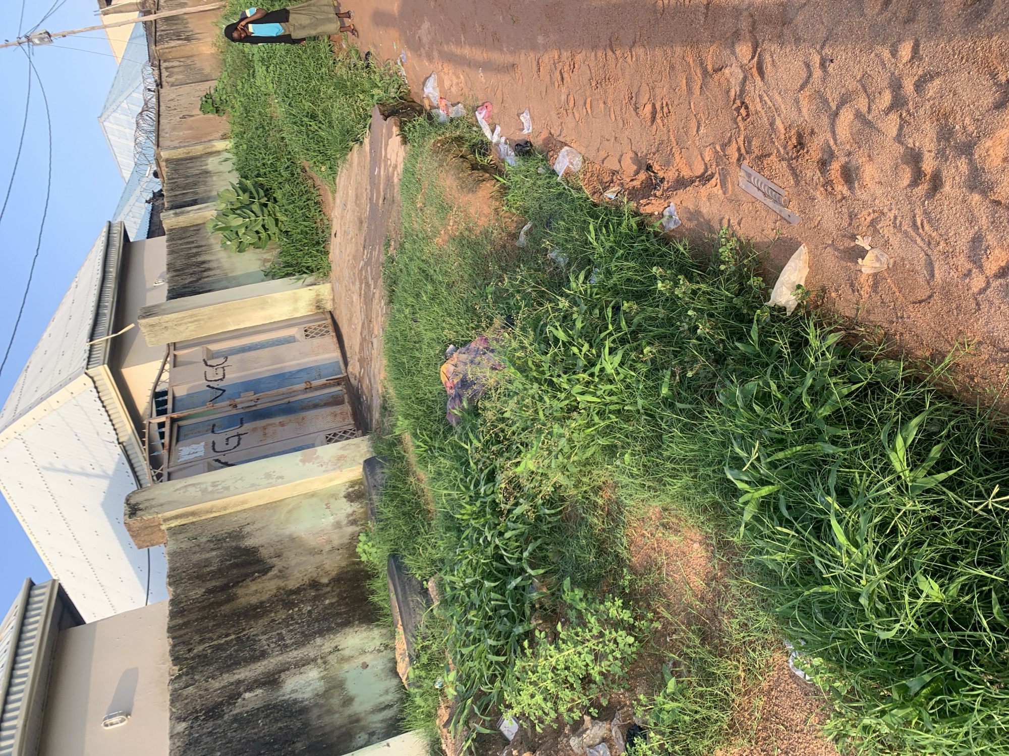 The image depicts a residential area in Jalingo, Nigeria. A concrete wall, stained with dark marks, encloses properties. Adjacent to the wall is a metal gate with faded blue and white paint, bearing graffiti that appears to read "VGC". The ground is a mix of sand and overgrown green grass, with some scattered debris including plastic bags, a torn piece of cloth, and a discarded tire. A person, dressed in a blue top and a long brown skirt, stands on the sandy path to the right, holding a phone to their ear. Further into the background, houses with corrugated metal roofs and barbed wire atop walls are visible. The sky is clear and blue, suggesting it is daytime.