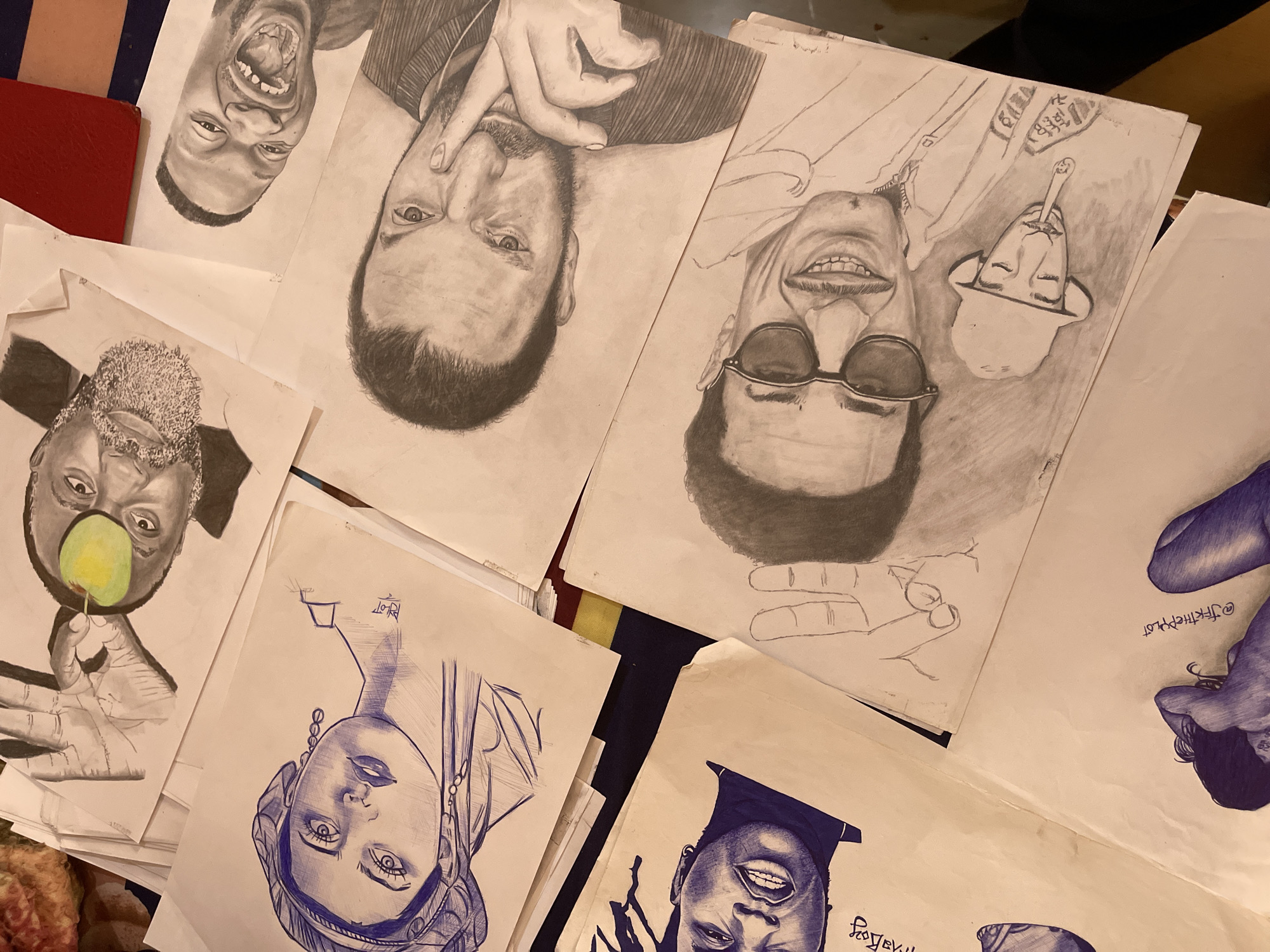 The image shows a collection of drawings scattered on a surface with a red book in the upper left corner. The drawings depict various portraits of people rendered in different styles using both pencil and pen. Some of the portraits are realistic, while others are more stylized. The scene appears to be indoors and the location is not visible. The image is focused on the drawings and there is no information to determine if this image was taken in Jos, Nigeria.
