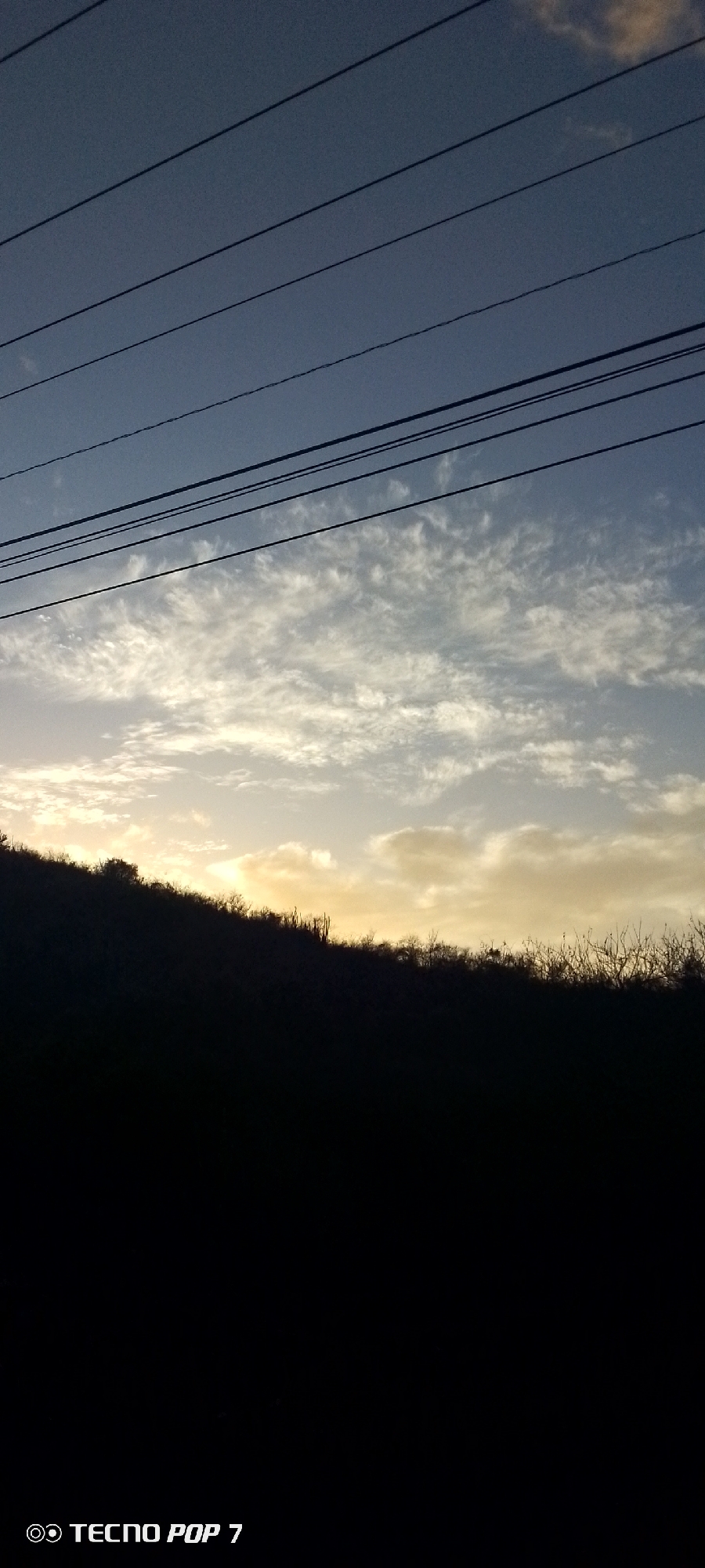 The image is a low-angle shot taken outdoors, likely near Altagracia, Venezuela. A dark, silhouetted hillside occupies the midground. A glimpse of foliage, including what appears to be a cactus, is visible on the hillside. The sky comprises the upper portion of the image, with a gradient from blue at the top to a yellowish-orange near the horizon, suggesting either sunrise or sunset. A layer of light, scattered clouds is visible. Several parallel power lines stretch across the upper part of the sky. The lower portion of the image is completely black, obscuring the foreground. The bottom left corner has the white text "TECNO POP 7", indicating the camera model used to take the picture.