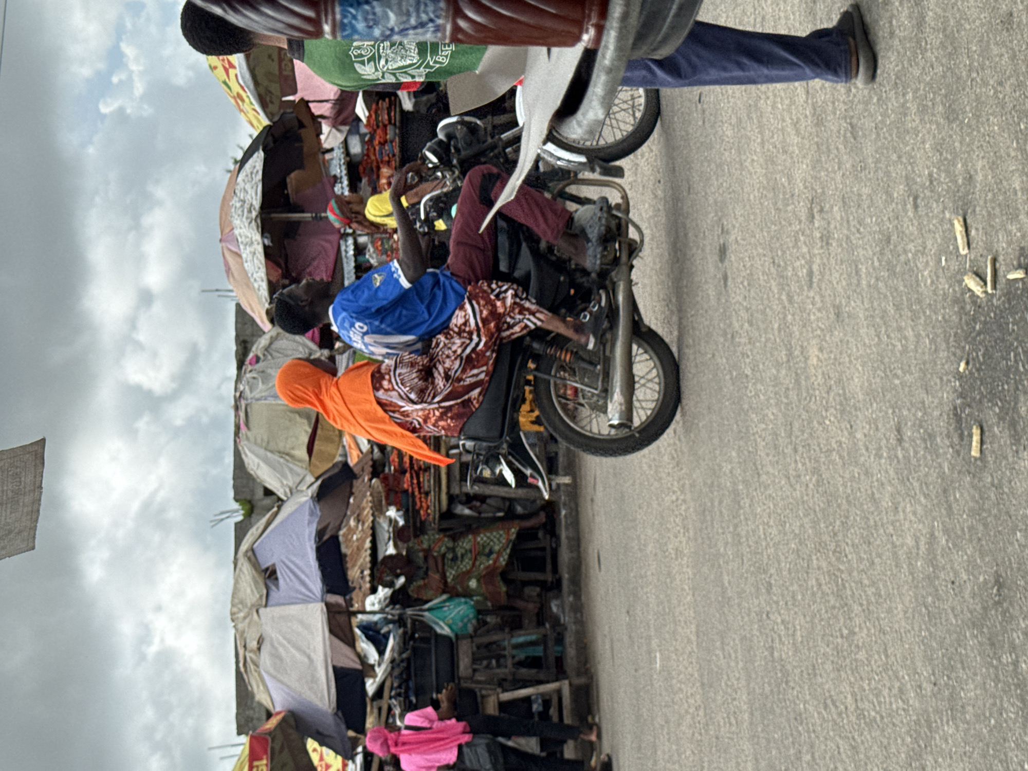 This is a vertical, full-shot photograph of a street scene, likely a marketplace, in Badagry, Nigeria. The dominant subjects are people and motorcycles. In the foreground, a person wearing a blue t-shirt and patterned leggings is seated on a motorcycle. They are holding onto the handlebars, appearing to be either riding or waiting. Behind them, another person in an orange headscarf and a patterned dress is also on the motorcycle. A third person, wearing a maroon long-sleeved shirt and dark pants, is also on the motorcycle, facing away from the camera.
The scene is filled with various objects. Motorcycles are a prominent feature, with one in the foreground and another, a classic style with a spoked wheel, partially visible to the right. There are also numerous market stalls, indicated by colorful umbrellas, stacks of fabric, and what appear to be goods displayed for sale. Some stalls are covered with tarps or corrugated metal. The ground is paved asphalt, with some debris scattered on it, including what look like discarded cigarette butts.
The sky is overcast with cloudy conditions, suggesting it could be any time of day but likely not bright sunshine. The overall atmosphere appears busy and perhaps a bit chaotic, typical of a bustling market environment. The people's expressions are not clearly discernible due to the distance and resolution, but their postures suggest engagement with the activity around them. There are no clear signs or text visible in the image. The combination of motorcycles, market stalls, and the people's attire strongly suggests a West African setting.