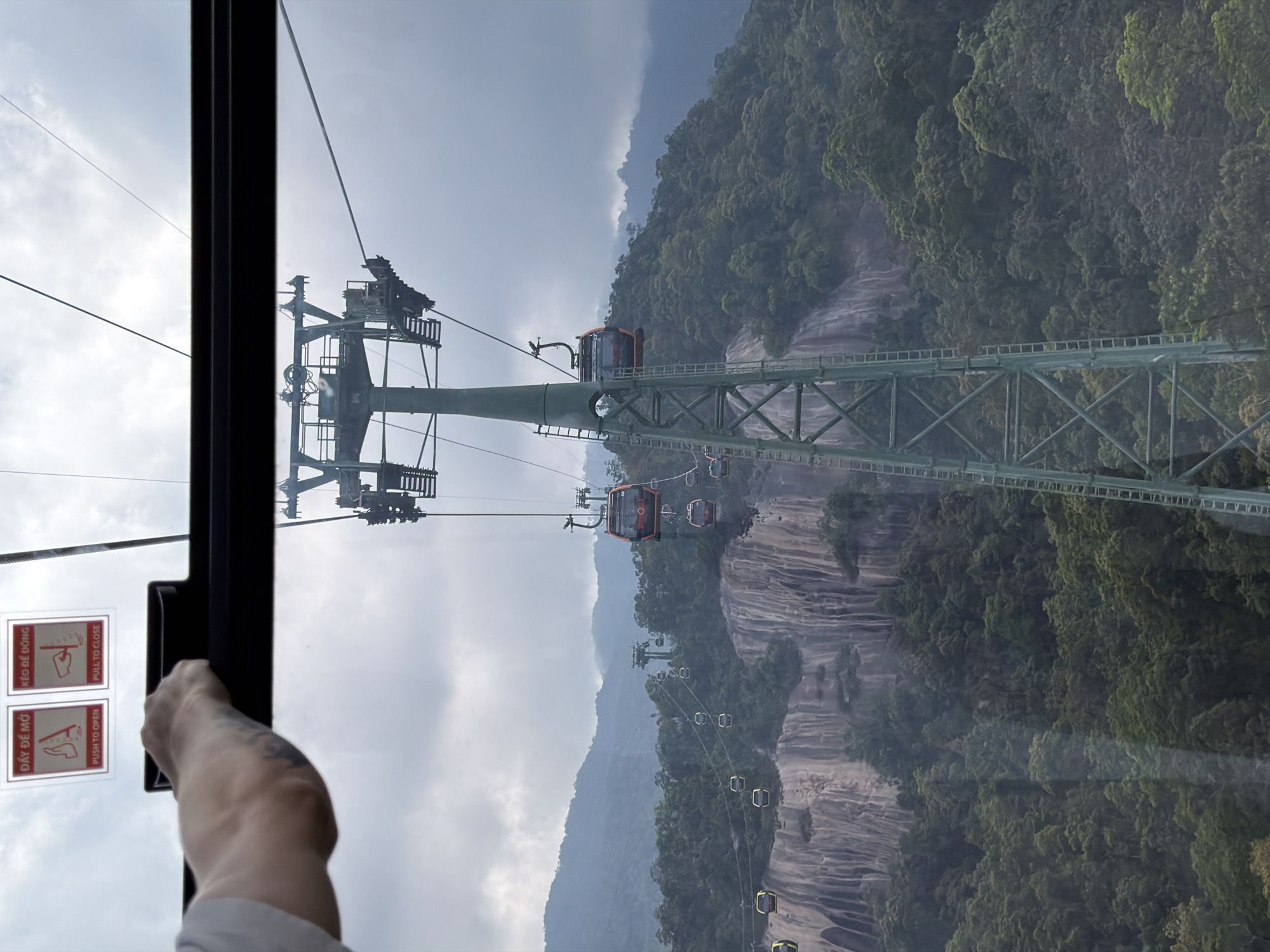 The image depicts a view from inside a cable car, looking out towards a mountainous landscape in Đà Nẵng, Vietnam. Visible through the window is a cable car support tower, and several other cable cars ascending or descending. Lush greenery and rock formations cover the mountainside. A hand reaches toward the cable car window frame, with visible tattoos on the forearm. The sky is overcast. Two warning signs are visible on the window.