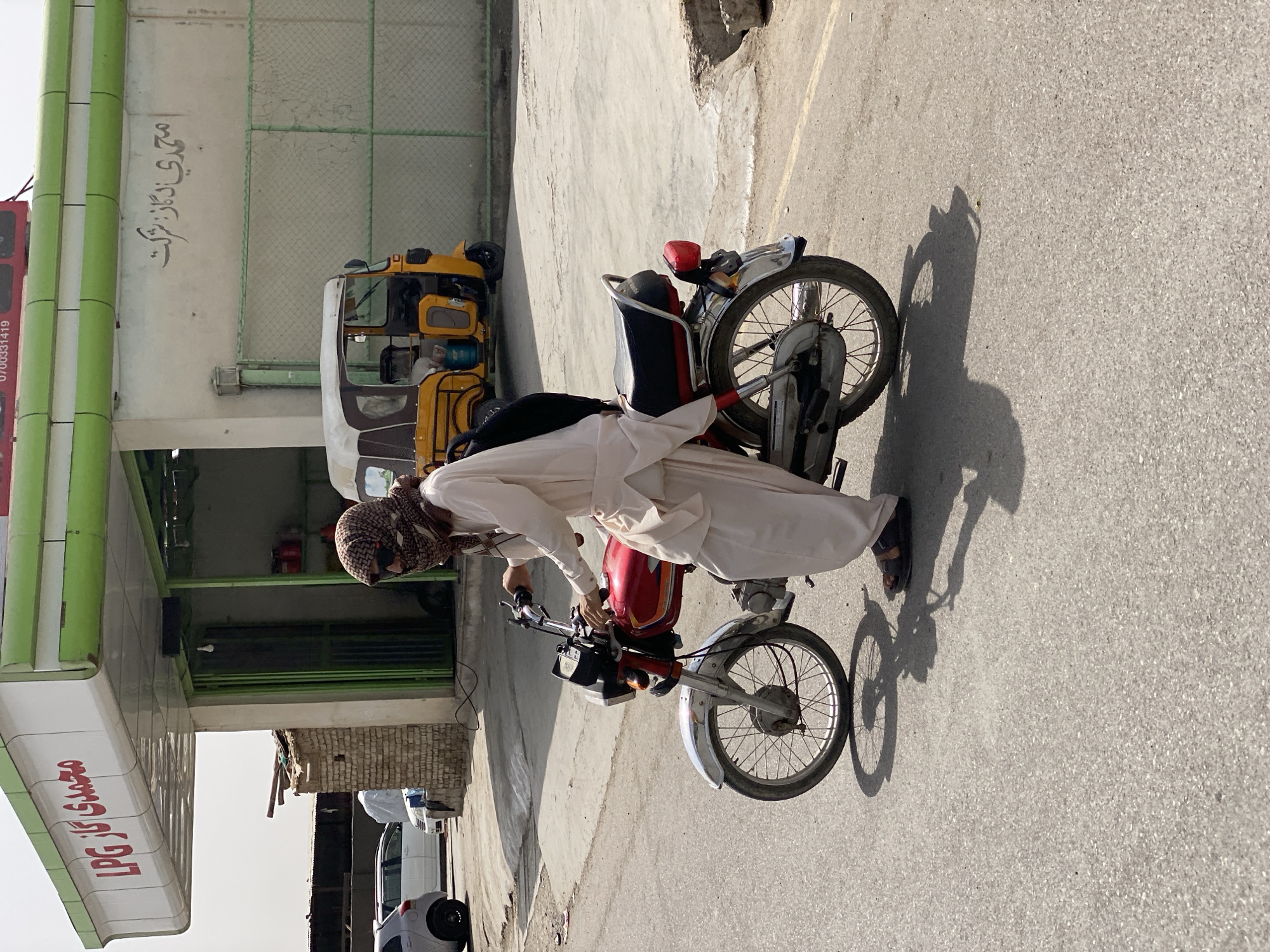 This is a full-shot, outdoor, eye-level, medium-light photograph of a man on a red motorcycle in a city environment. The man is wearing a light-colored shalwar kameez and a patterned headscarf that covers his face, along with sunglasses. He is on a street, with a gas station in the background. The gas station has a green awning with the text "محمدي گاز LPG" in red. Behind him, a yellow auto-rickshaw is parked. The ground is asphalt, and the sky is overcast. The overall impression is that of a typical street scene in Kandahar, Afghanistan.