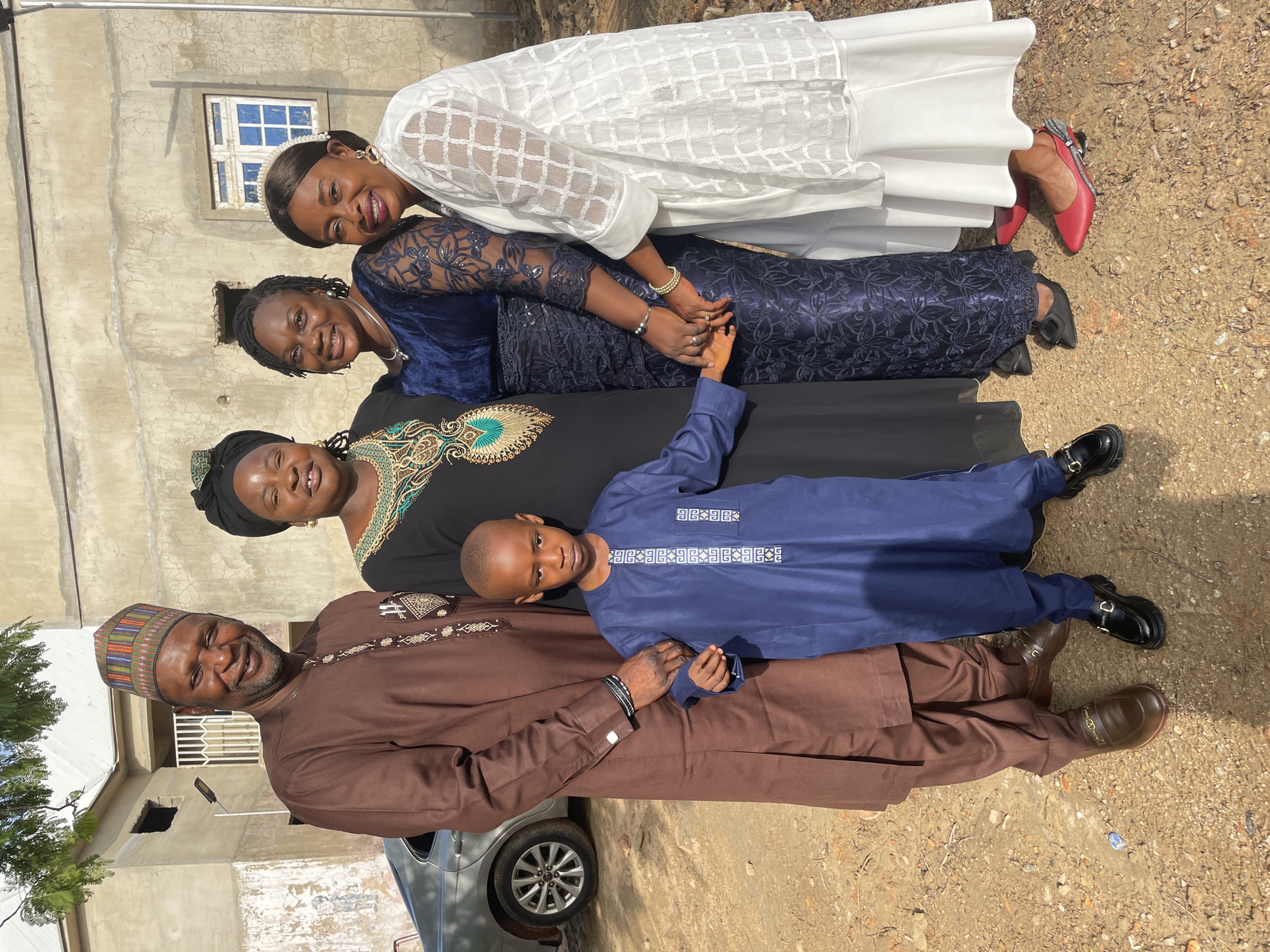 In an outdoor shot in Akwanga, Nigeria, a group of five individuals—four women and one boy—are horizontally posed against the backdrop of a partially visible silver car and a building with a window. A man in a brown traditional attire and multicolored cap stands at the leftmost position, and a boy in a blue outfit is positioned above him. Above the boy, there is a woman wearing a black dress, complemented by a green and gold embroidered neckline and a black headscarf. Another woman above is dressed in a navy blue lace dress. A final woman at the top wears a white dress and red shoes. The people in the picture are holding hands. The ground is a dirt surface.