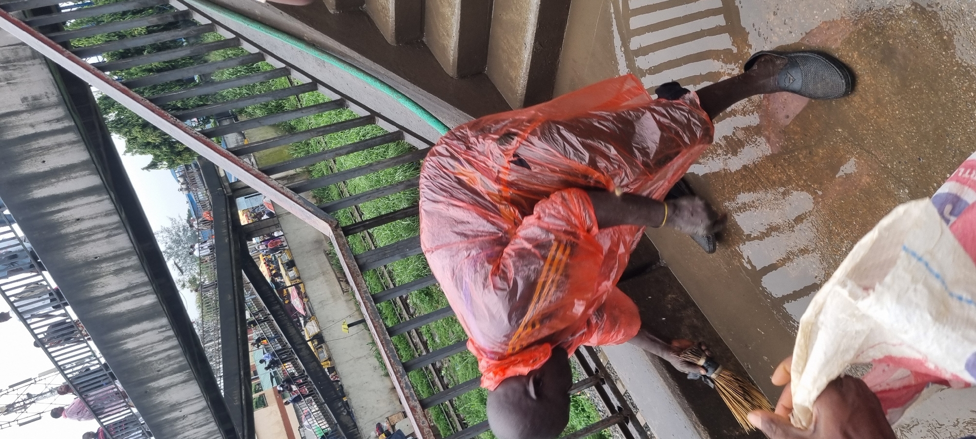 In Ikeja, Nigeria, a person wearing a bright orange poncho is sweeping water off a concrete surface with a broom. The surface appears to be a walkway or elevated path, with a railing and a set of stairs visible. Beyond the railing, train tracks with overgrown grass are present. In the background, an overhead pedestrian bridge is crowded with people and vehicles, indicating a busy urban environment. The overall scene suggests an outdoor public space that is being maintained.