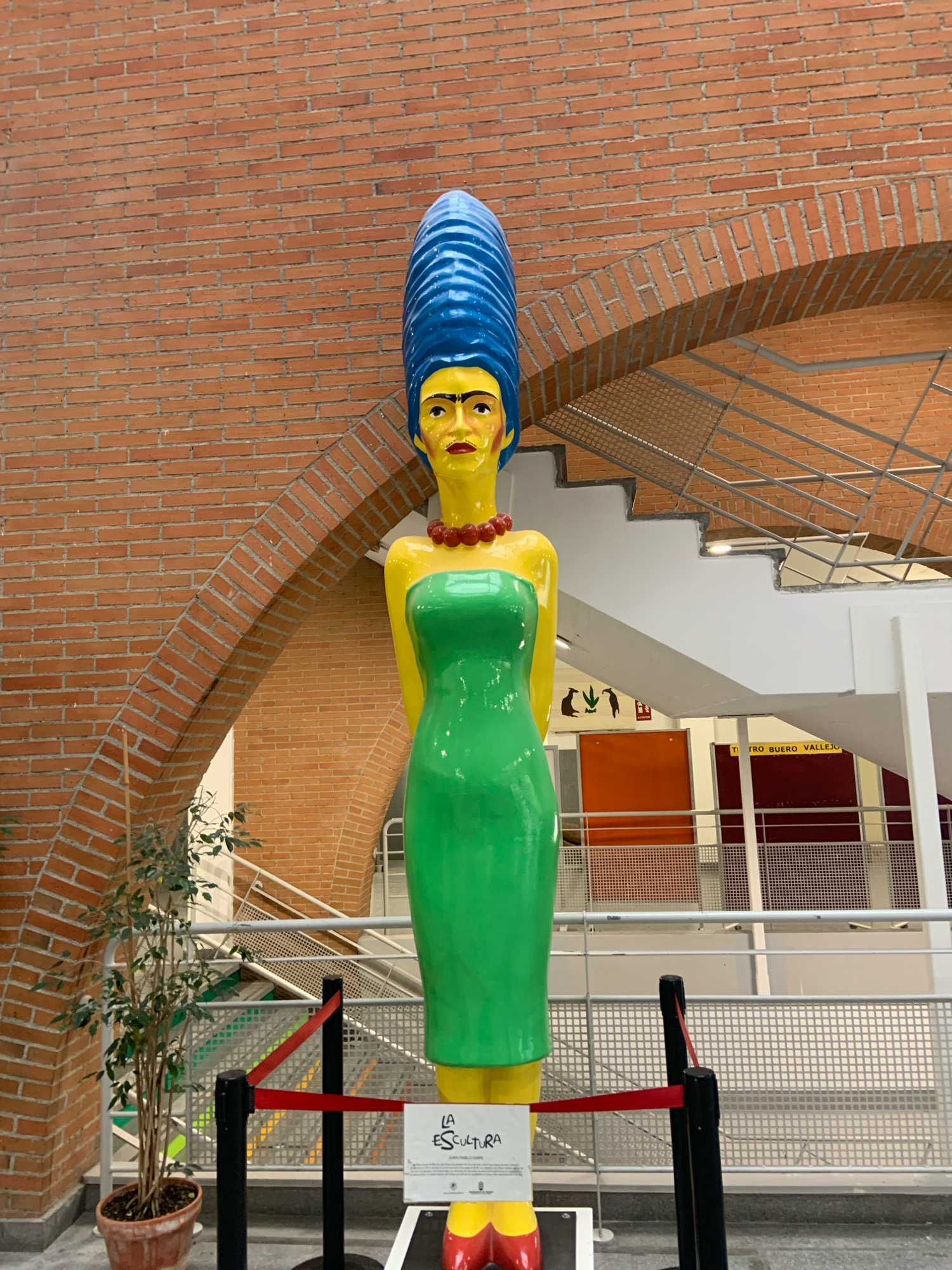 A large, colorful sculpture of a woman with an exaggerated blue beehive hairstyle, yellow face, and green dress stands prominently in the foreground. The sculpture is positioned in front of a textured brick wall with an arched opening. To the left of the sculpture, a potted plant with thin branches and green leaves is visible. Behind the sculpture, a staircase with metal railings ascends, and various signs and colored panels are mounted on the wall. A red rope barrier with black stanchions is placed in front of the sculpture, indicating a designated viewing area. The overall environment suggests an indoor public space, possibly an exhibition hall or cultural center. Based on available information, this sculpture is likely located at the Centro Cultural Viñamar, in Alcorcón, Spain, as part of an exhibition or permanent display.
