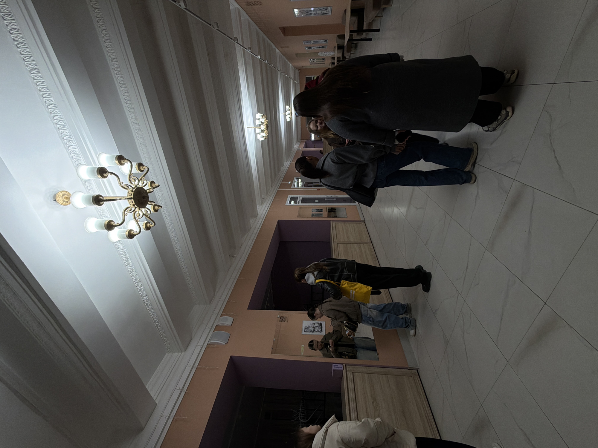 A dimly lit hallway in what appears to be a public building in Perm, Russia, possibly a university or cultural center. The hall is long and narrow, with tall ceilings adorned with ornate plasterwork and chandeliers. Several people are present. A group of three individuals are standing closer to the camera, appearing to be engaged in conversation. One is wearing a dark coat and jeans, another a gray jacket and jeans, and the third is wearing a brown jacket and blue jeans. A person wearing a yellow bag is slightly further away from the camera. The hall has a light-colored, tiled floor, and the walls are painted in peach and purple colors. A mirror reflecting the surroundings is visible on the right.
