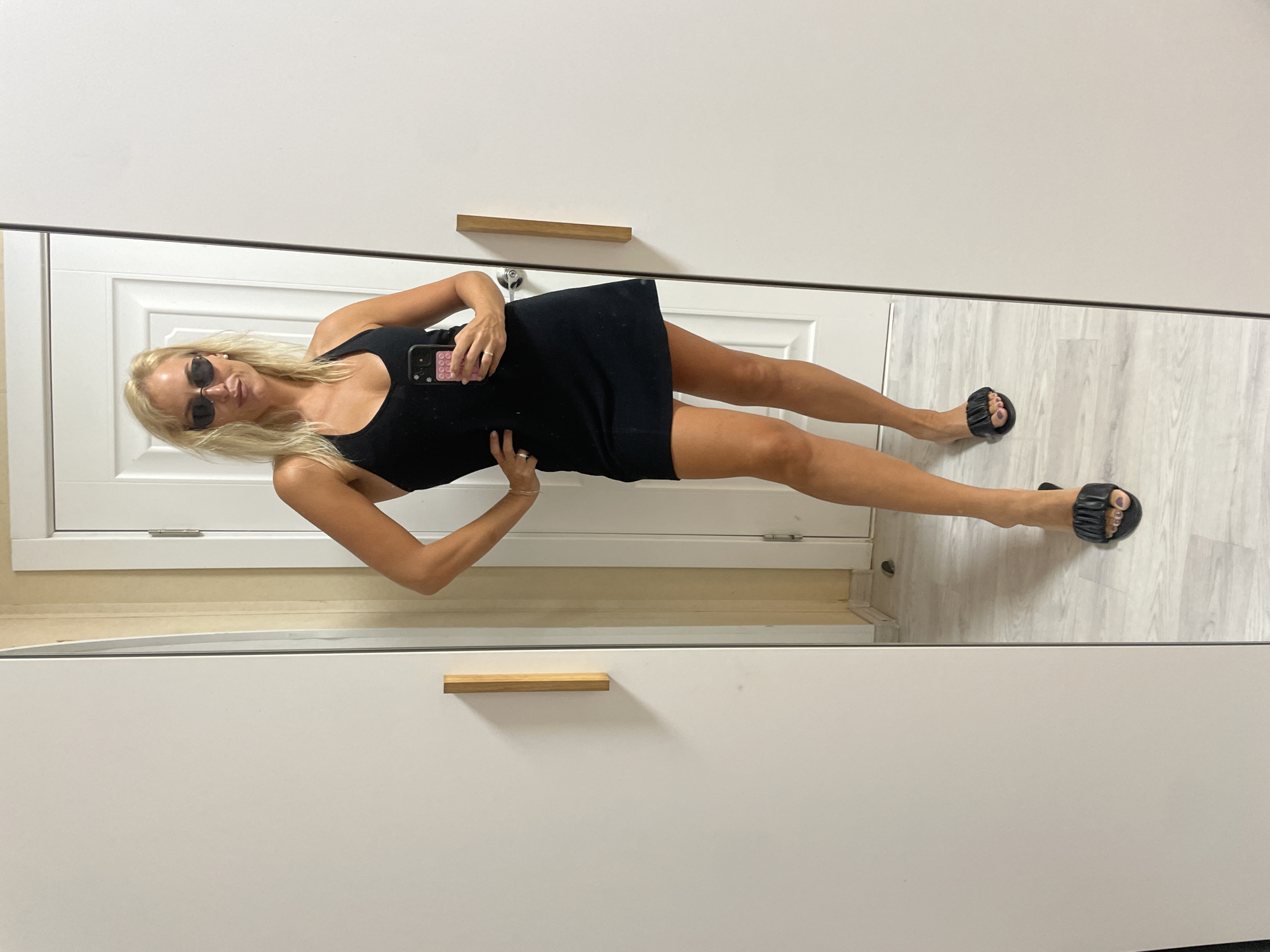 A blonde woman stands in a mirrored room, taking a selfie. She is wearing a black dress, black slide sandals, and dark sunglasses. Her phone is visible in her right hand. The room appears to be a bedroom or dressing room, with a large white wardrobe with wooden handles. The flooring is light-colored wood. The lighting suggests it is daytime. There is no visible text in the image.