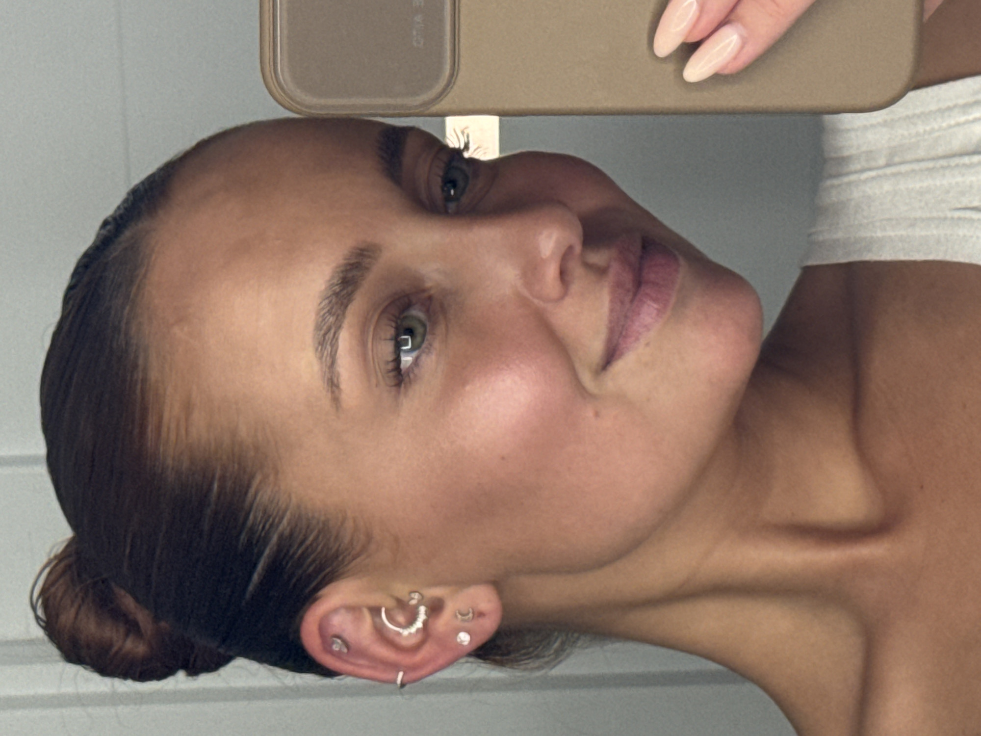 The image shows a close-up, profile view of a young woman with brown hair pulled back in a bun. She has green eyes and fair skin with subtle freckles. She is wearing multiple earrings in her left ear, including a hoop with small stones and moon-shaped studs. The woman is holding a tan smartphone in her right hand, angled to capture her reflection. Her nails are painted a pale pink. She is wearing a light-colored ribbed top. The background appears to be an indoor setting with a neutral-colored wall. There is no identifiable information linking the image to the city of Chaikovsky, Russia.
