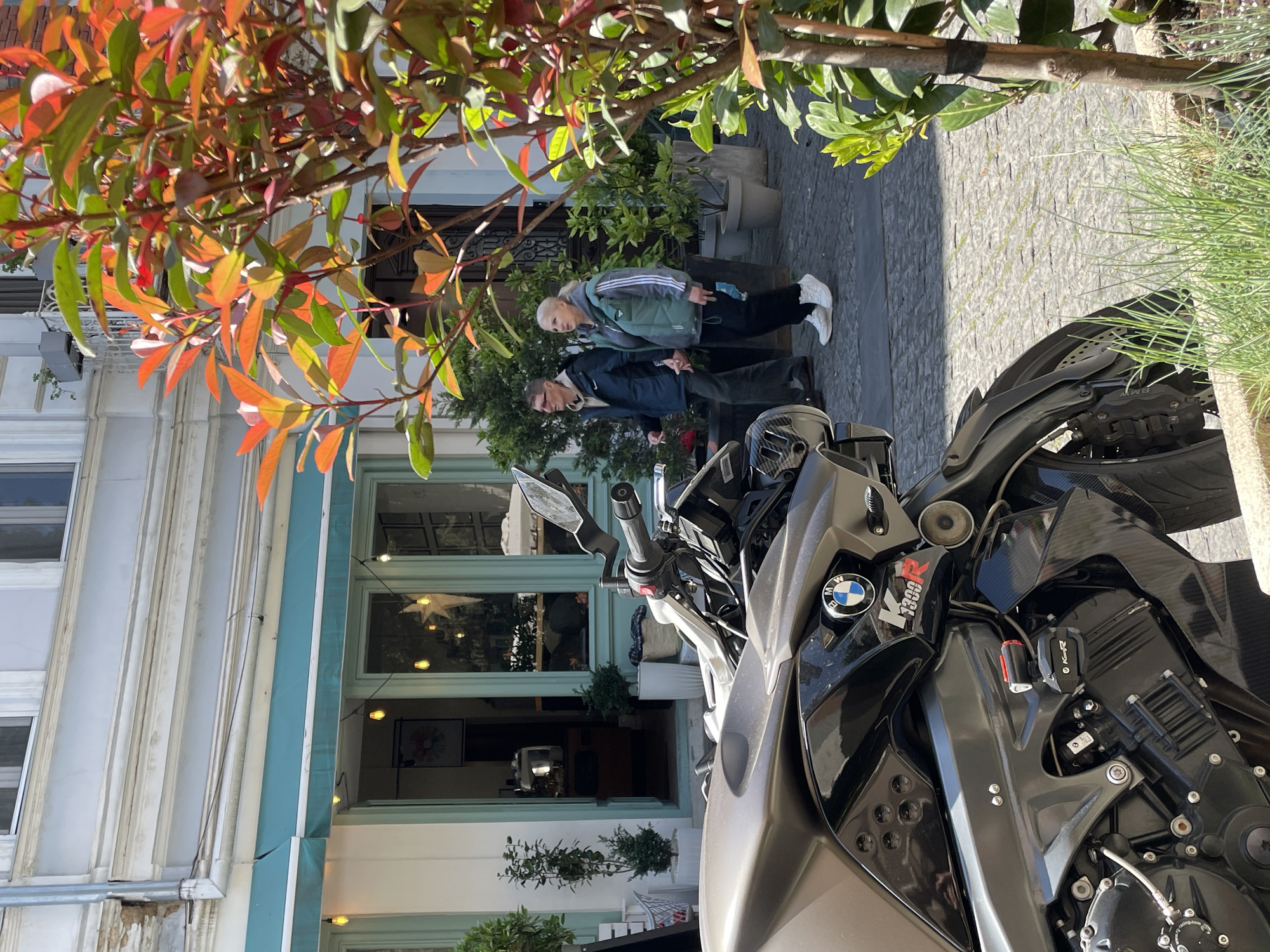 The image captures a vibrant street scene in Tbilisi, Georgia, under bright daytime sunlight. In the immediate foreground, a grey and black BMW K 1300 R motorcycle is prominently featured, with its front fairing, handlebars, and part of the engine visible. Visible text on the motorcycle includes the "BMW" logo, "K 1300 R," and "BMW Racing."
Two individuals are seen standing on a cobblestone sidewalk behind the motorcycle. An older woman with light hair is wearing a green Adidas track jacket, black pants, and white sneakers. She appears to be looking towards the left side of the frame. Slightly in front of her is an older man, possibly wearing glasses, dressed in a dark blue Nike jacket and dark pants, facing towards the right.
The background reveals a light-colored building with large windows and a blue-teal awning or trim, suggesting a shop or cafe. Decorative string lights are visible through the windows. The scene is lush with greenery, including trees with bright orange-red and green leaves in the upper left, and various potted plants lining the sidewalk. The ground is paved with light-colored cobblestones, indicating an urban setting. The clear, sunny weather casts distinct shadows, suggesting a bright day, possibly late morning or afternoon. The people appear to be casually standing or strolling, contributing to a relaxed everyday street ambiance.