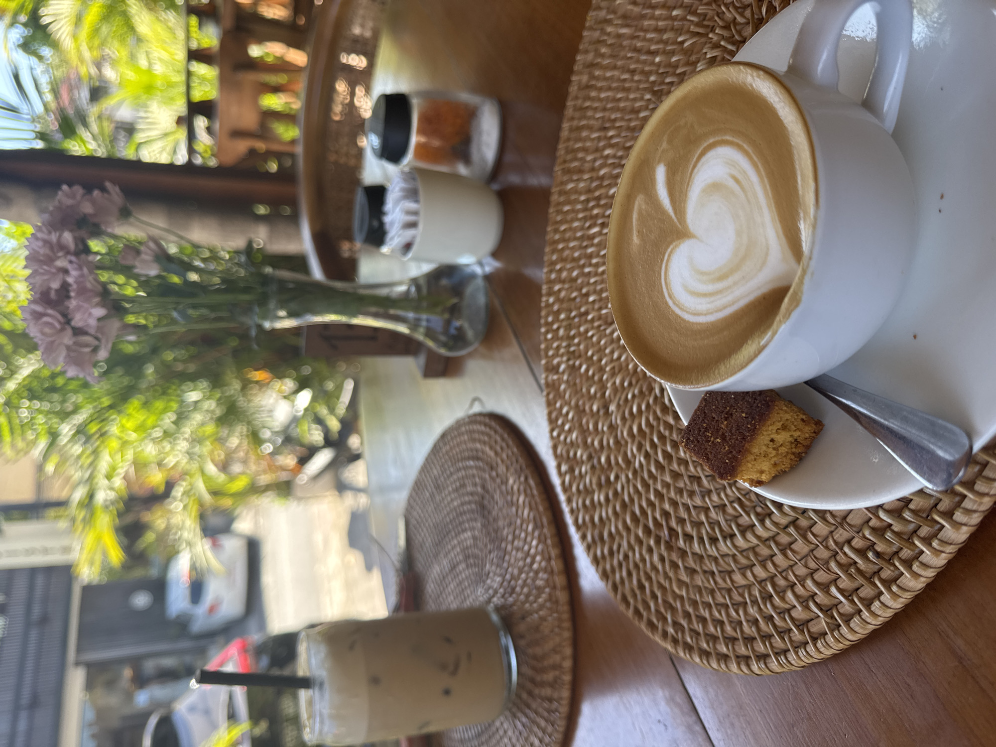 The scene is a cafe or restaurant setting, likely outdoors due to the foliage visible through the window. A cup of coffee with latte art in the shape of a heart rests on a saucer, accompanied by a small piece of cake. The cup and saucer are placed on a woven placemat. To the left, there is a vase with flowers and a glass of iced coffee with a straw. Salt and pepper shakers, along with a container of toothpicks, are placed on the table near the coffee. The environment suggests a relaxed, casual atmosphere.