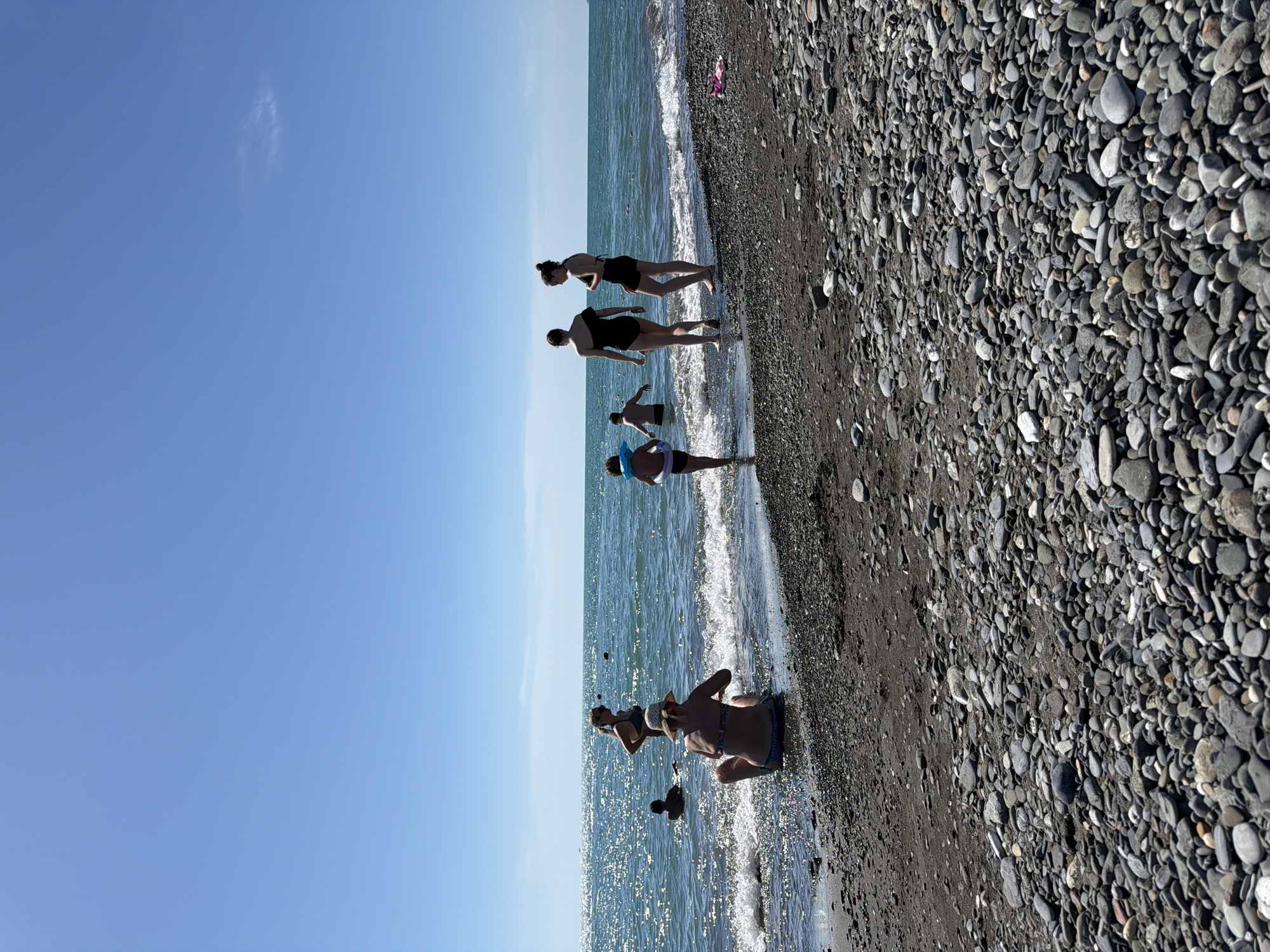 In Sochi, Russia, on a sunny day, a group of people are enjoying the Black Sea coast. The beach is covered in pebbles. In the foreground, a woman wearing a bikini and a straw hat sits on the shore, facing away from the camera. Further back, several individuals are in the shallow water, with some wading and others swimming. Two women in black swimsuits stand near the water's edge, while a child with a blue inflatable ring around their waist is entering the sea. The water is calm and reflects the sunlight, creating a shimmering effect. The sky is a clear, bright blue with a few wispy clouds.